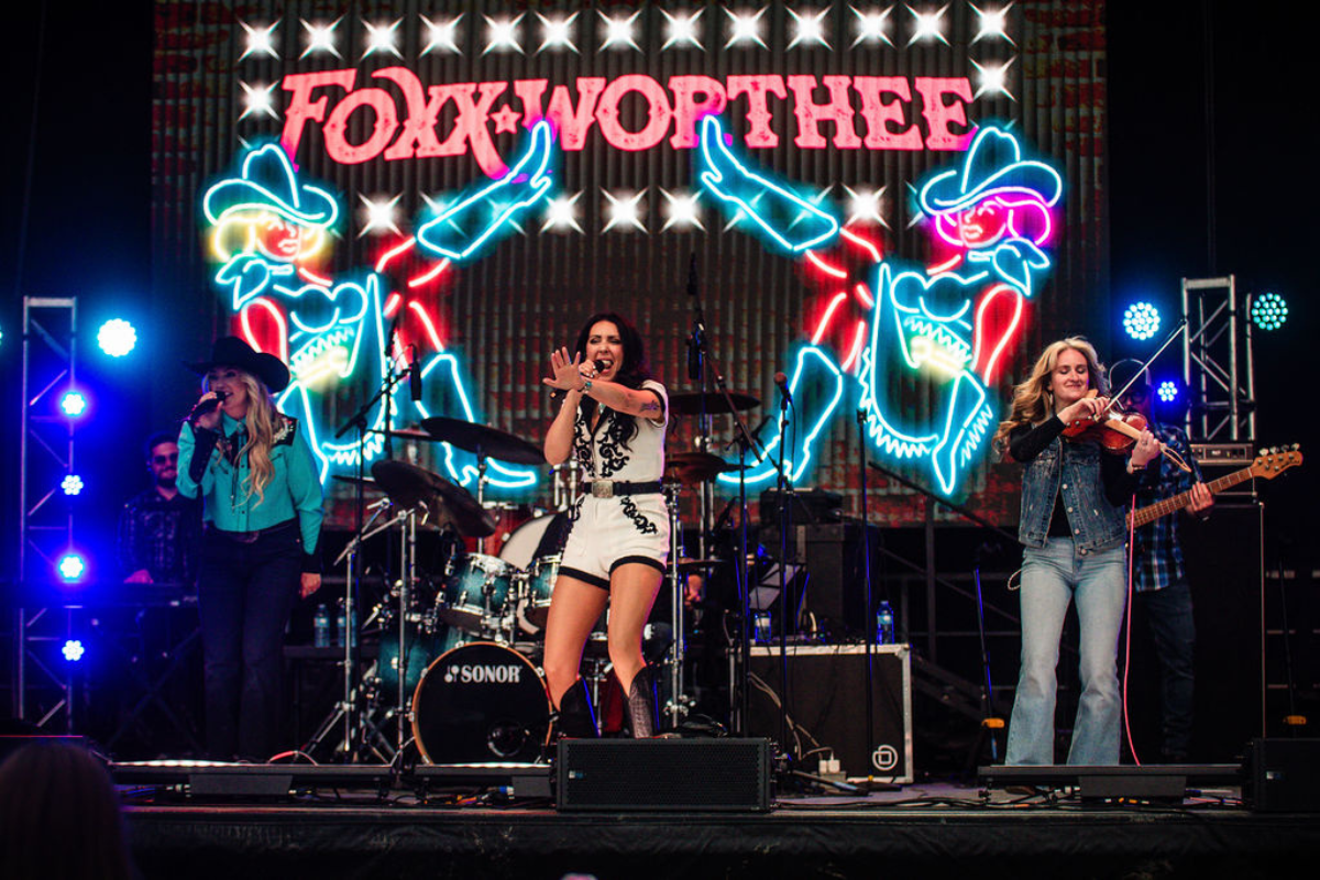 Foxx Worthee on stage with neon lights behind. Two singers and a fiddle player with a backing band.