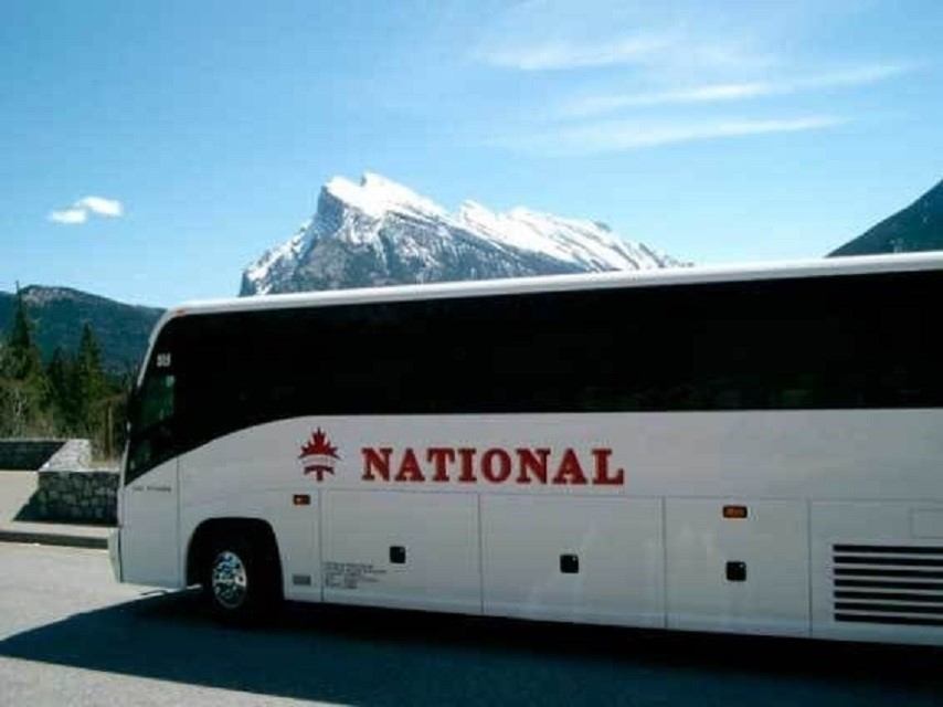White coach bus with 'NATIONAL' logo parked near snowy mountains under blue sky.