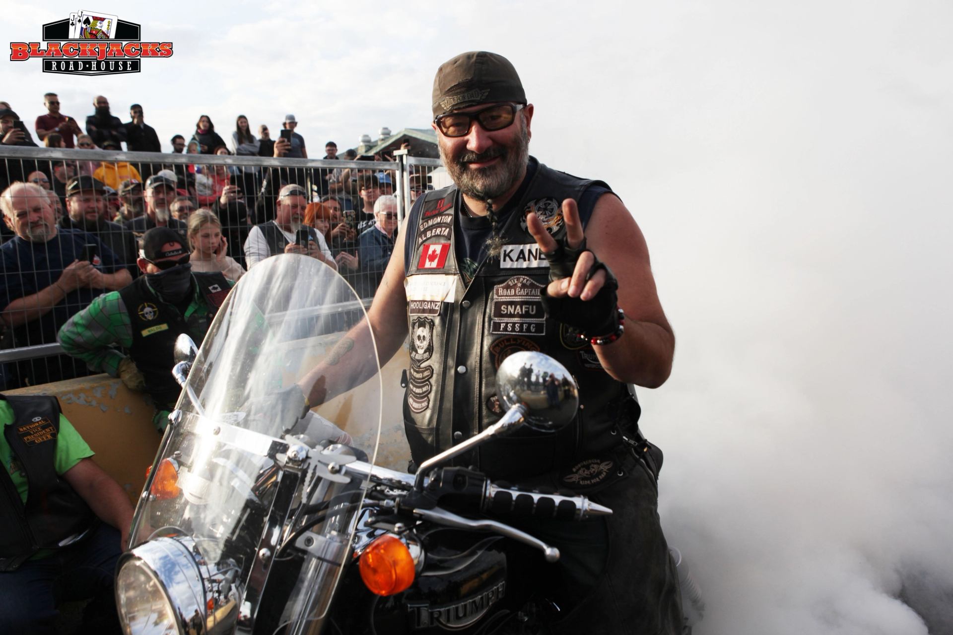 Motorcyclist in black vest rides, pointing with hand, crowd behind metal fence and smoke visible.