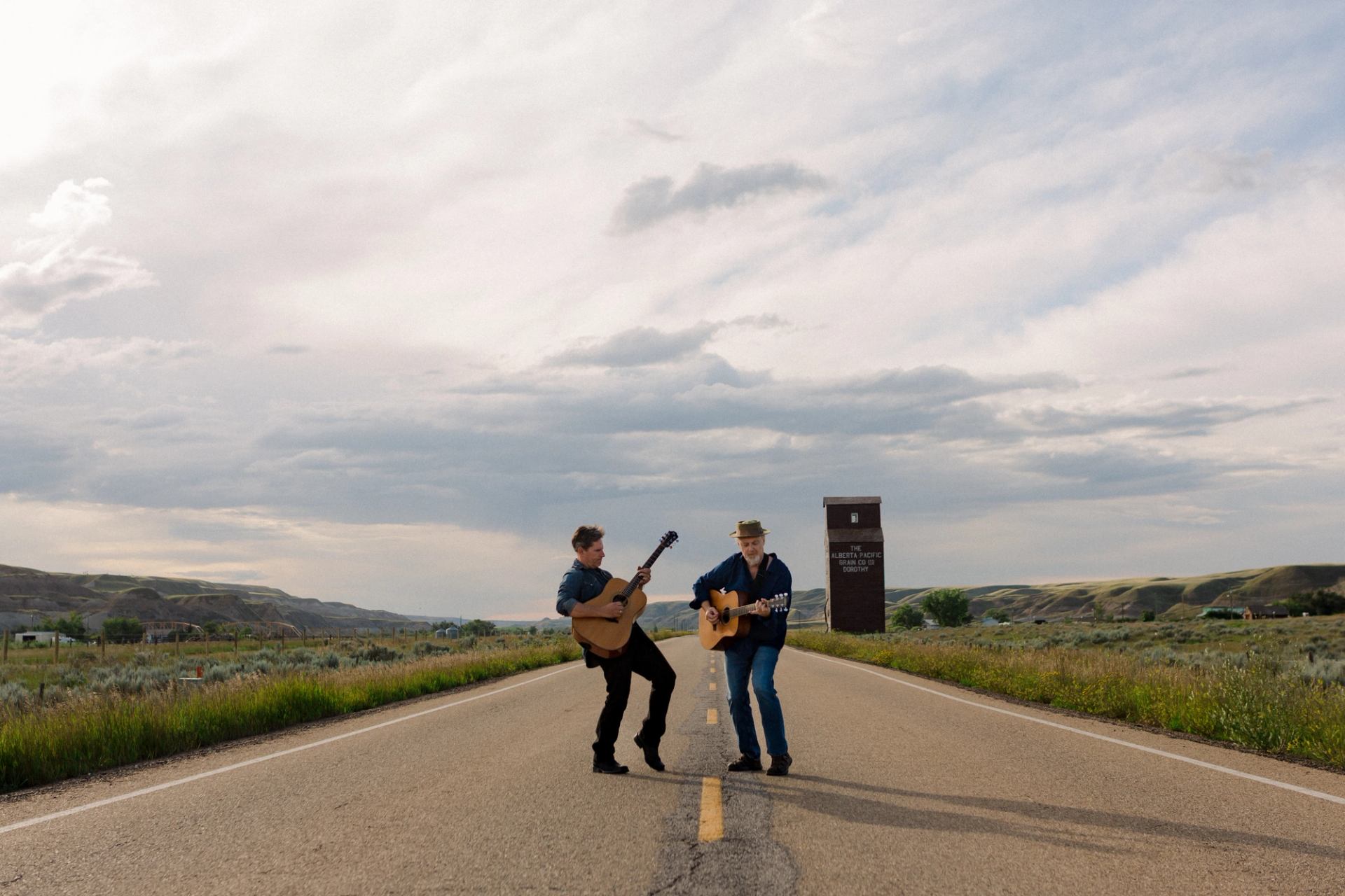 Two people play acoustic guitars on a rural highway with open fields and hills.