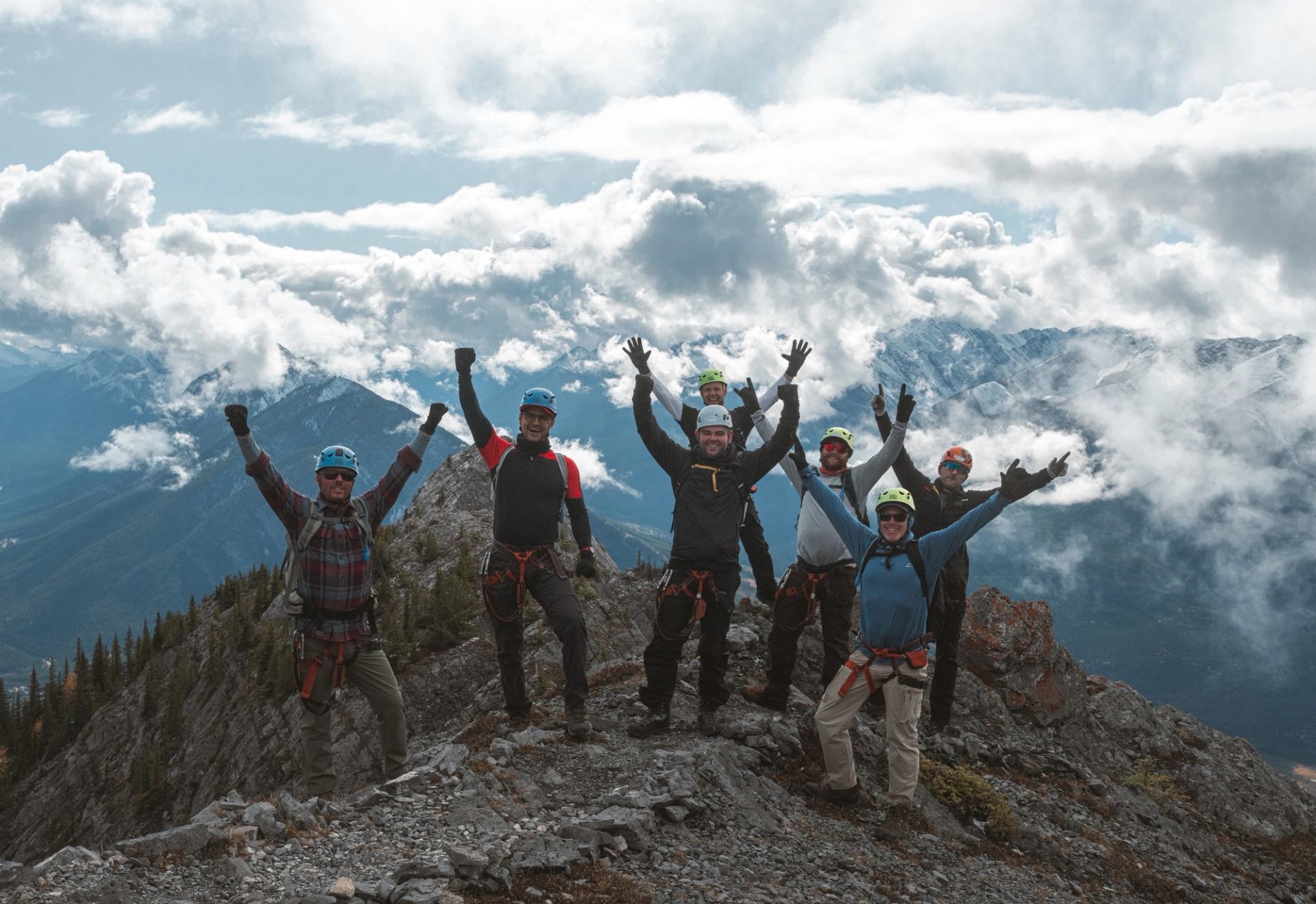 Hikers with arms raised on a mountain summit with clouds and peaks in the background.