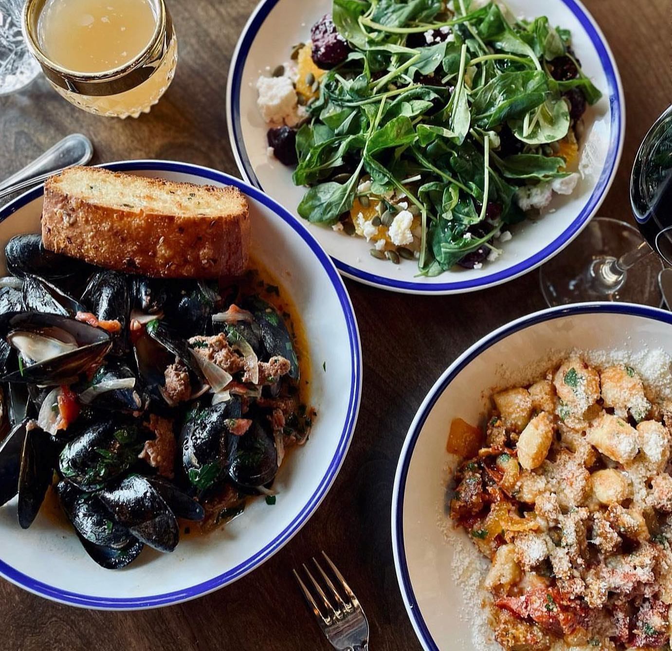 Three plates with salad, mussels and bread, and gnocchi pasta on a wooden table with drinks.
