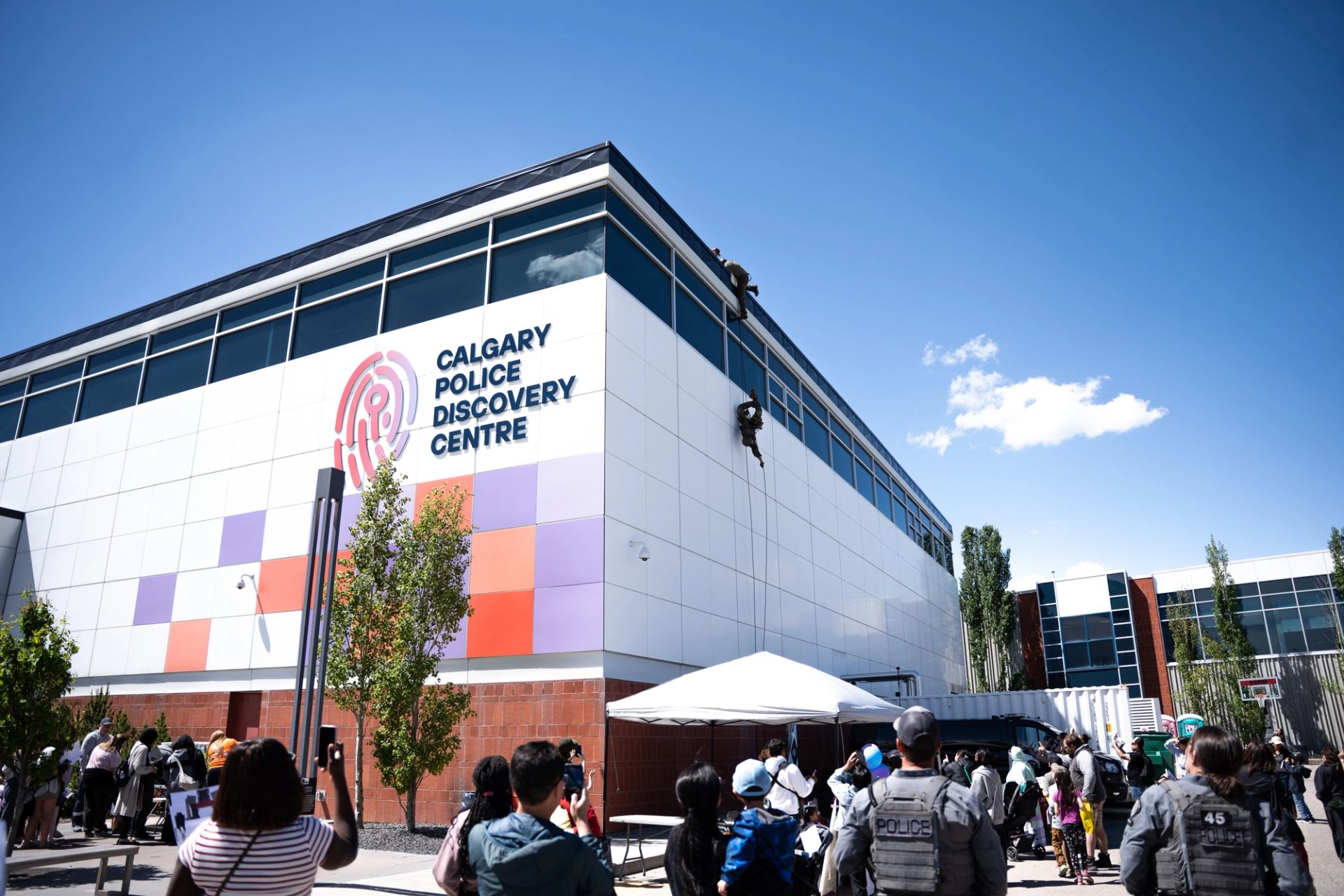 Crowd watches police officer rappel down Calgary Police Discovery Centre building exterior.