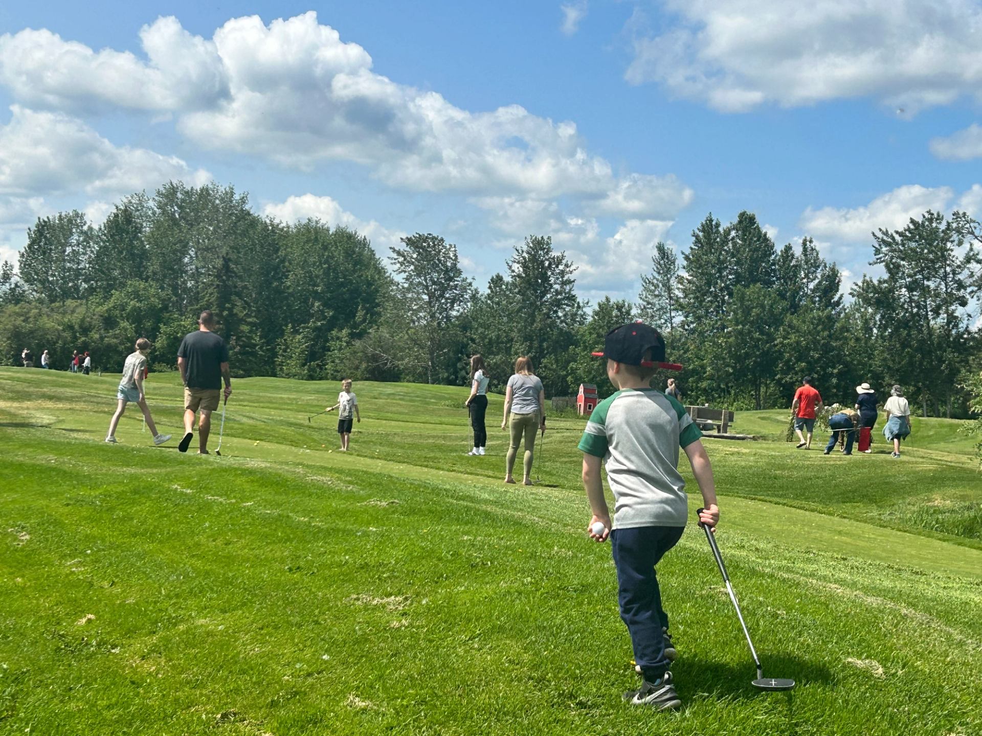 Mini golf players walk on a grassy course under blue sky and clouds.