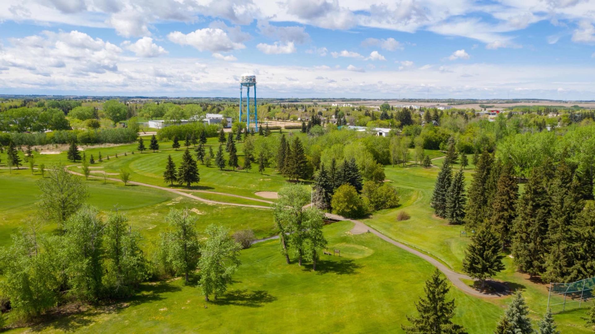A wide shot of a golf course with a water tower and trees, under a bright sky.