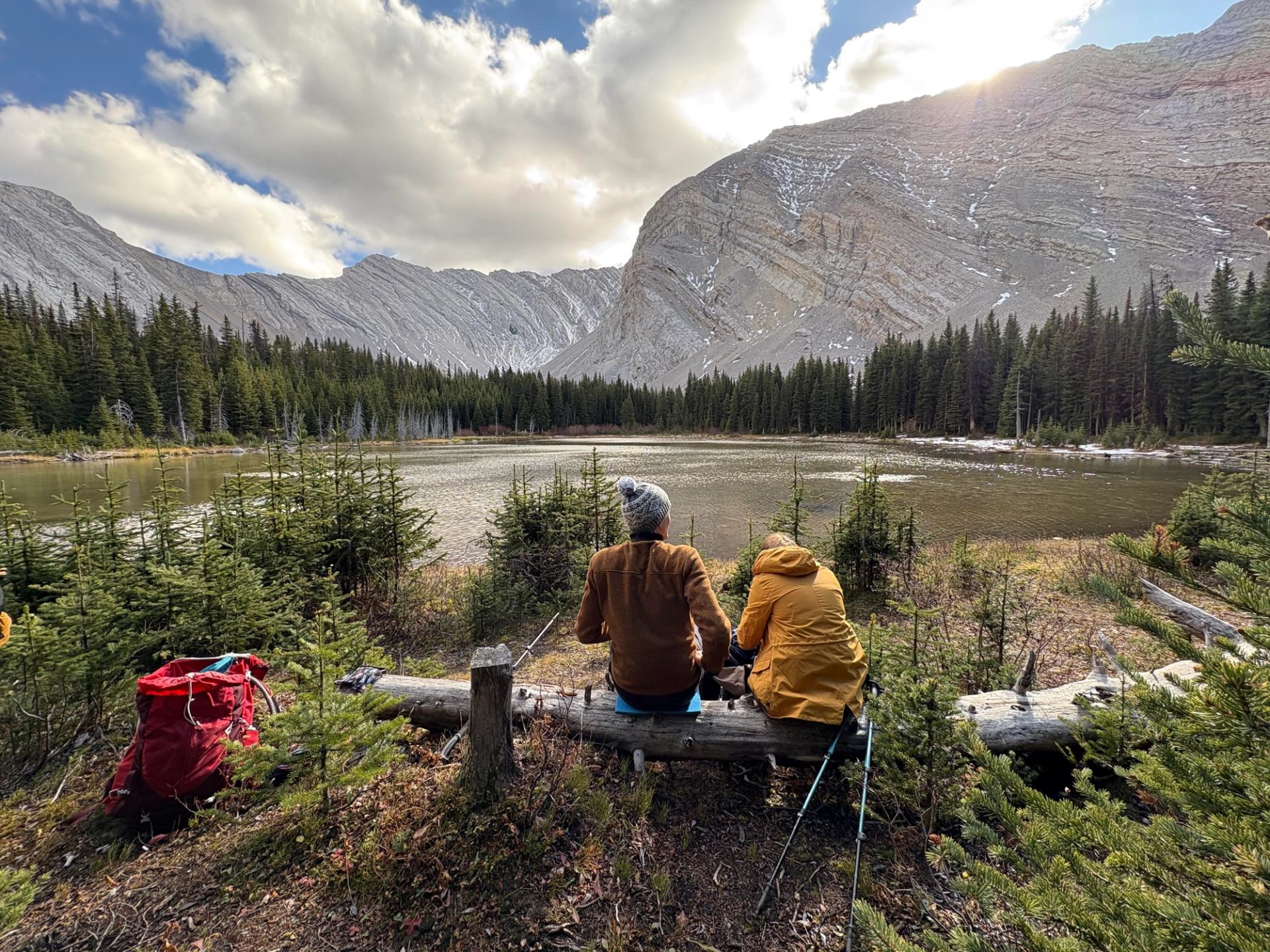 Two hikers resting on a log by a calm alpine lake surrounded by forest and rocky peaks.