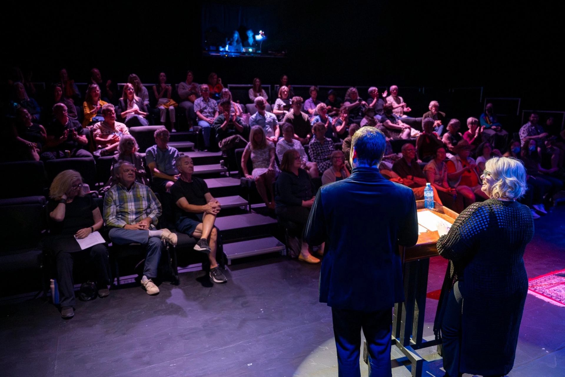 Audience sits in tiered theatre seating while two speakers stand at a lit podium.