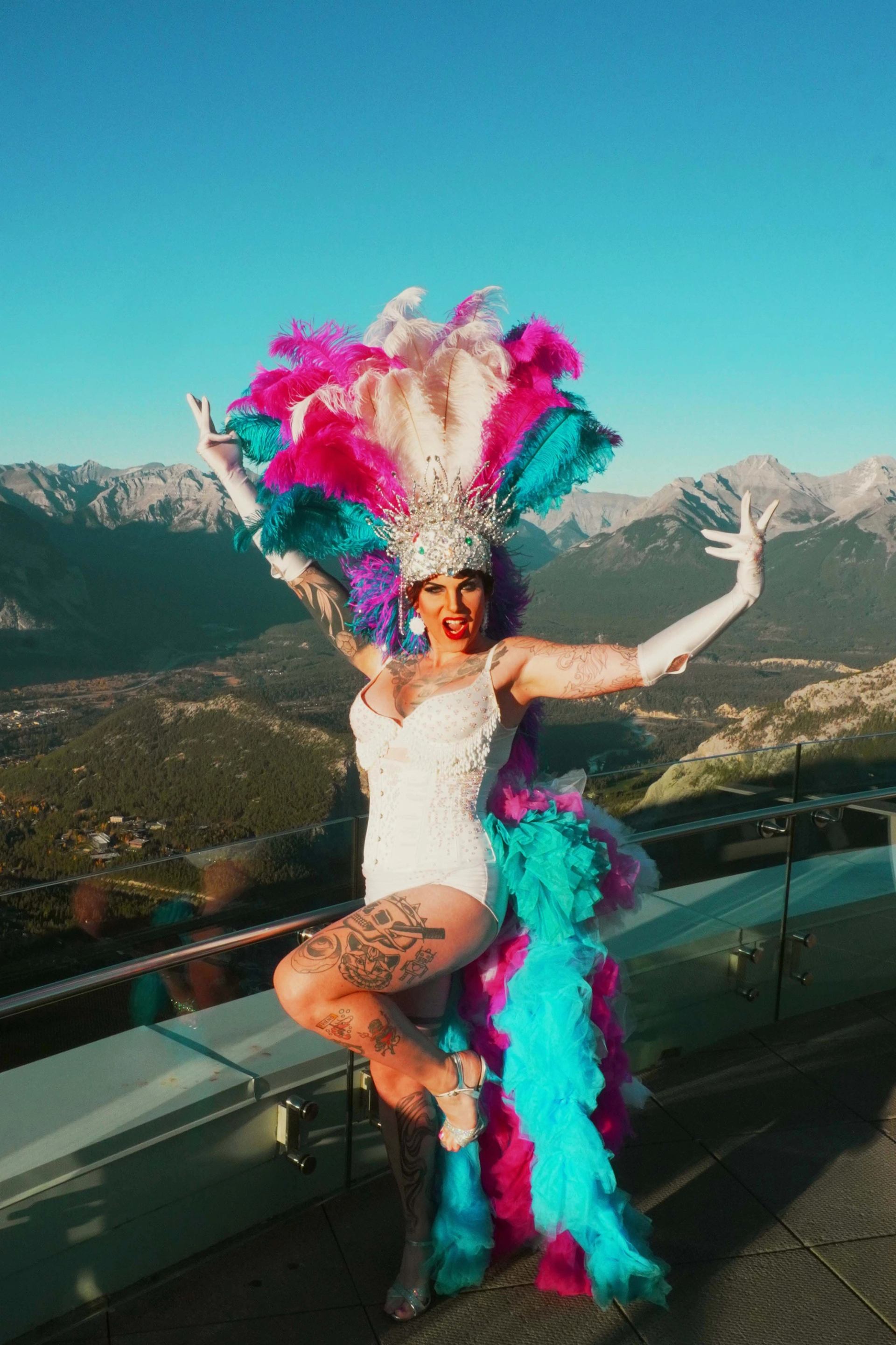 A drag performer in a sparkling bodysuit and dramatic feathered headdress poses confidently on a mountain viewpoint, with a vast alpine landscape in the background.