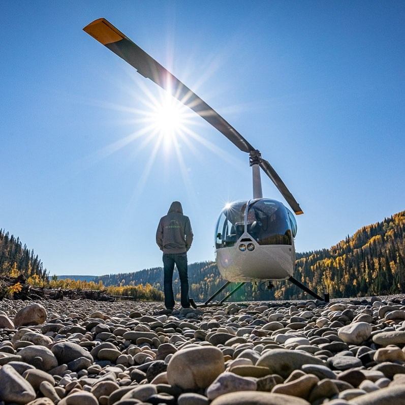 Helicopter on rocky terrain with person nearby and autumn trees in background.