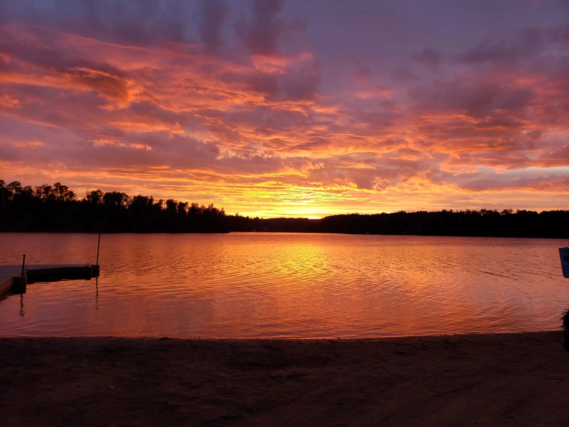 Lake at dusk with rippling water, shoreline, and trees beneath colorful clouds.