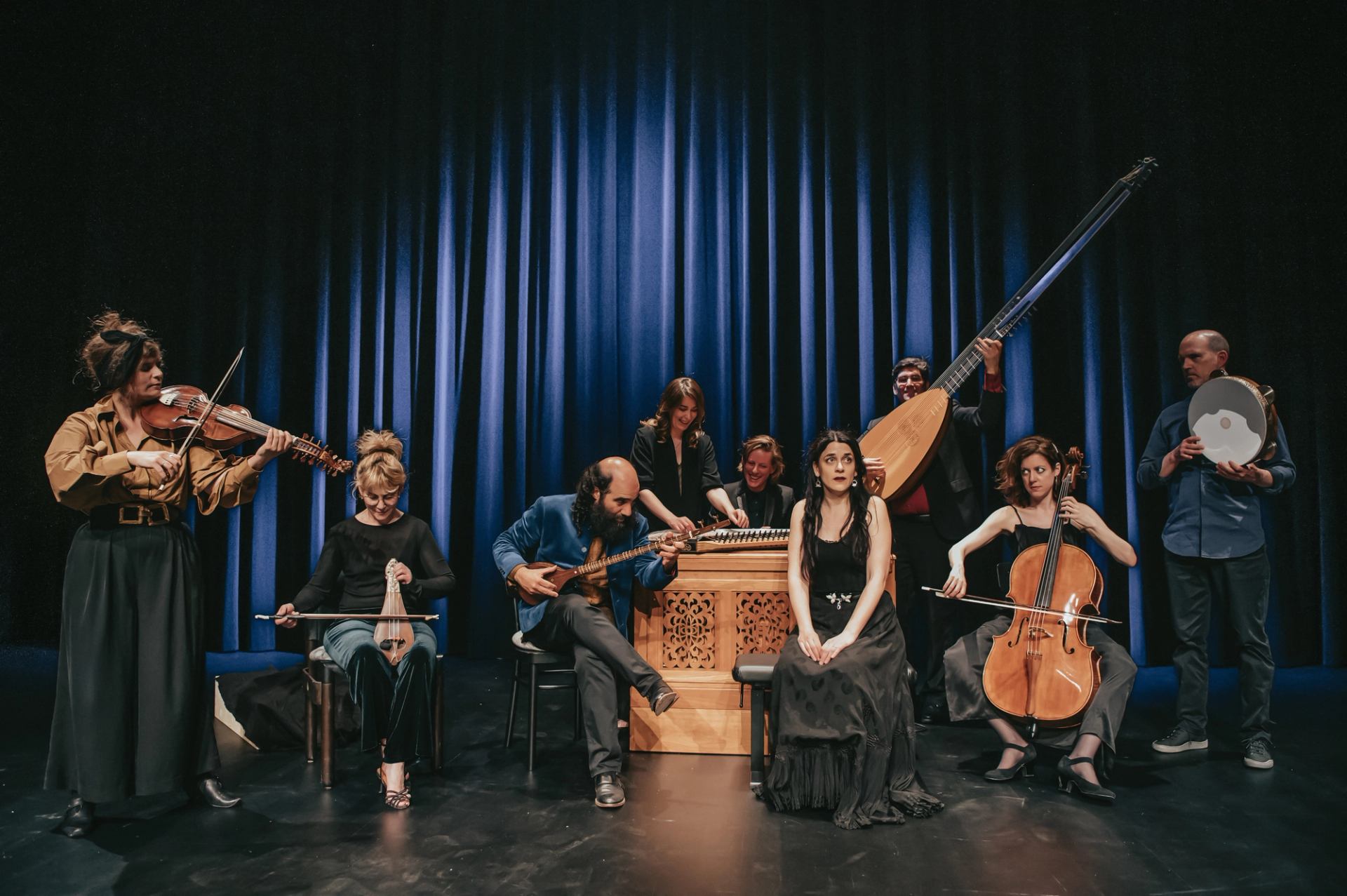 Ensemble of musicians onstage with string and percussion instruments before blue curtains.