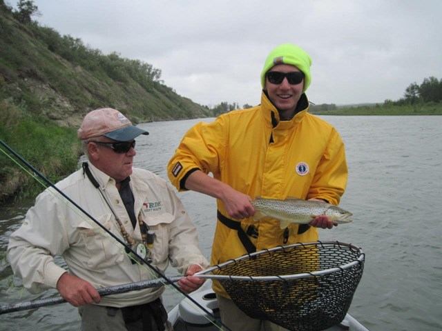 An older man holds a fishing net, while a younger man holds a fish next to it.