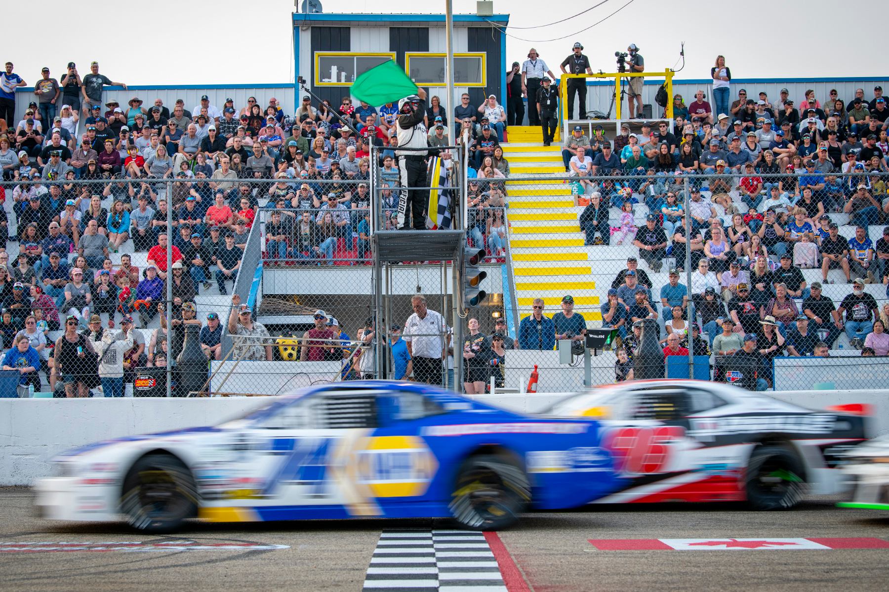 Two NASCAR cars speed past the start line as the green flag waves with full grandstands behind.