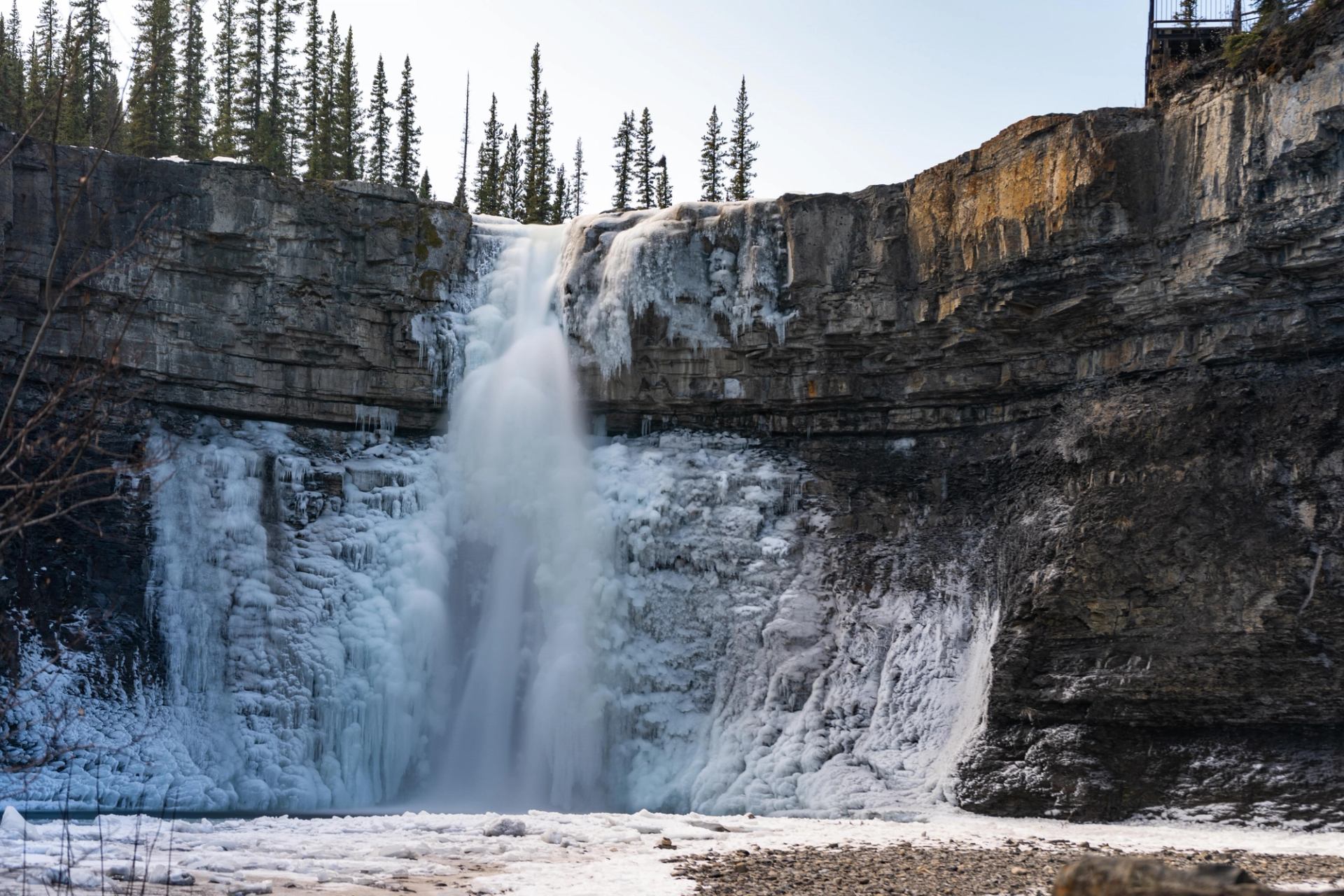 Waterfall plunging over rocky cliffs with pine trees in background, surrounded by snow and ice.