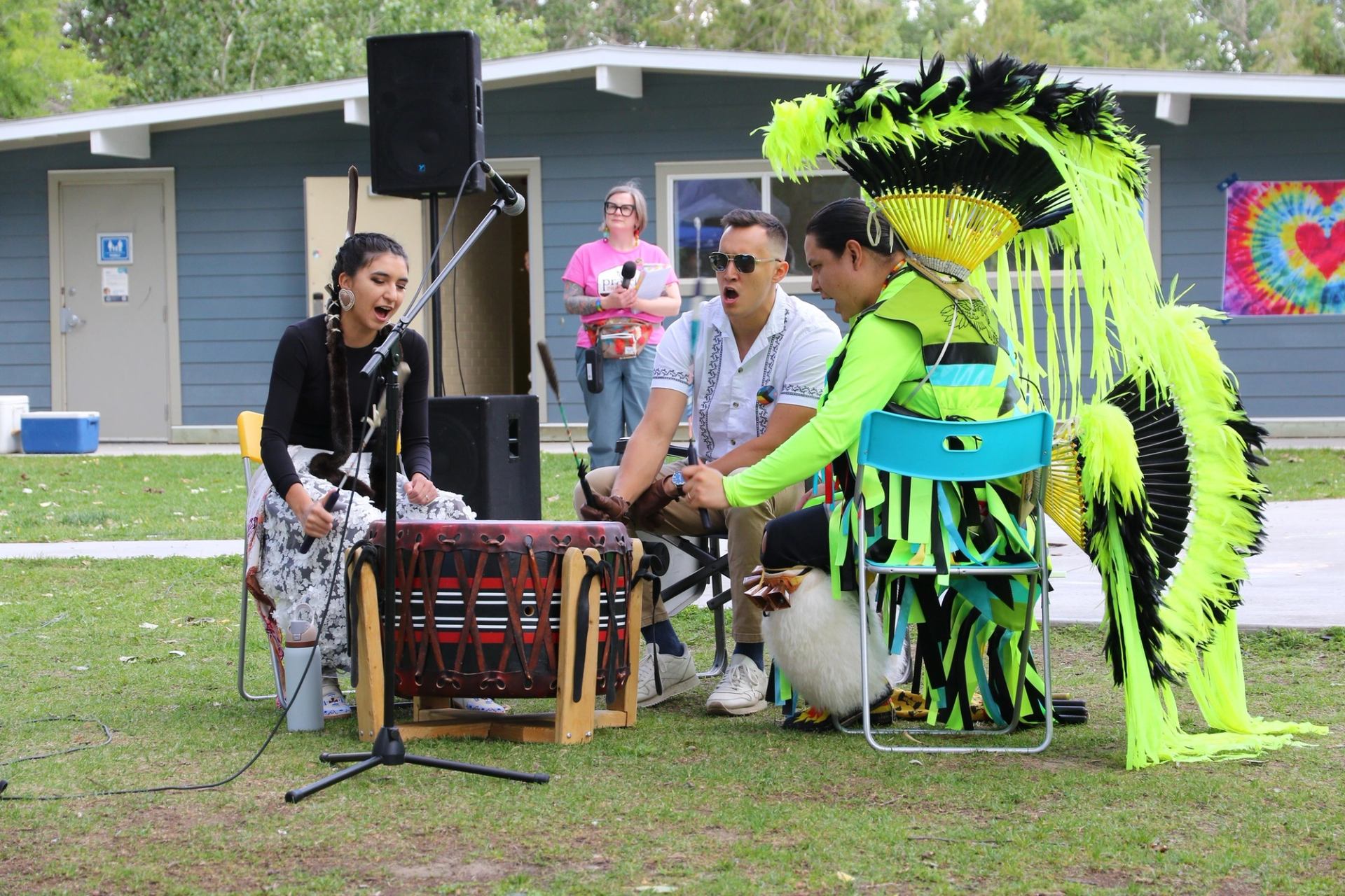 Drummers and singer perform outdoors, one wearing bright regalia with large feather bustle.