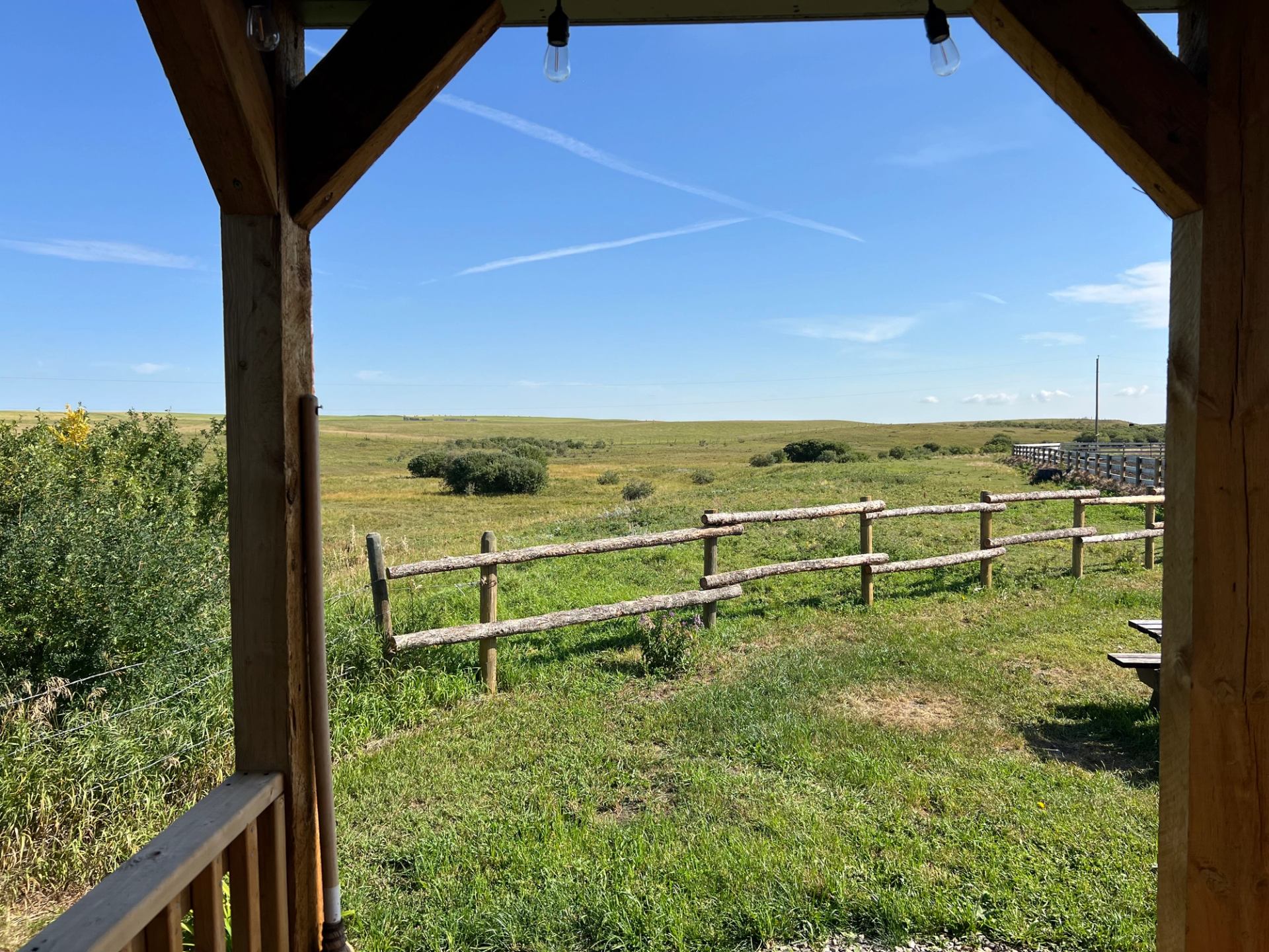 View from a cabin porch overlooking green fields and a wooden fence under a clear blue sky.
