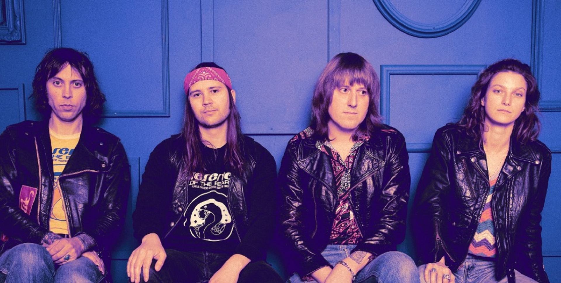 Four musicians in casual wear sit on a bench against a blue wall.