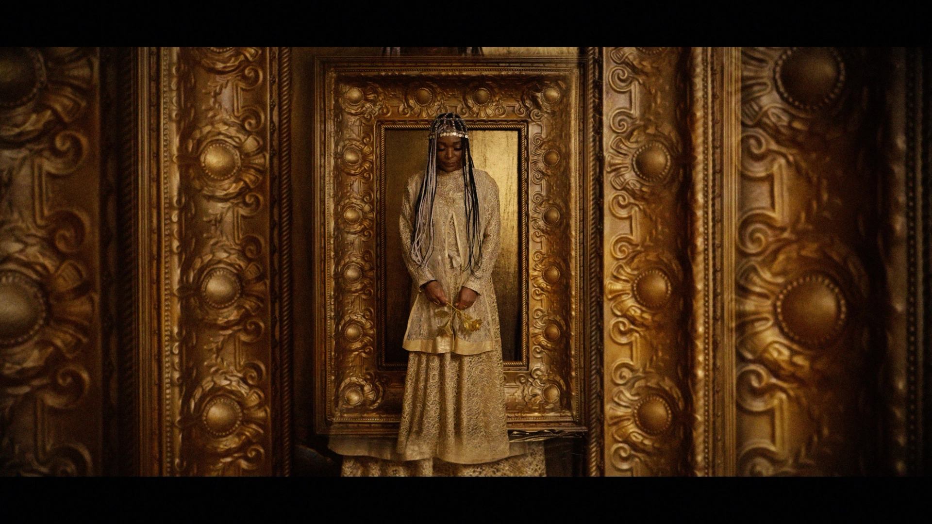 Person in gold robe stands against ornate gold-framed wall, holding small bouquet.