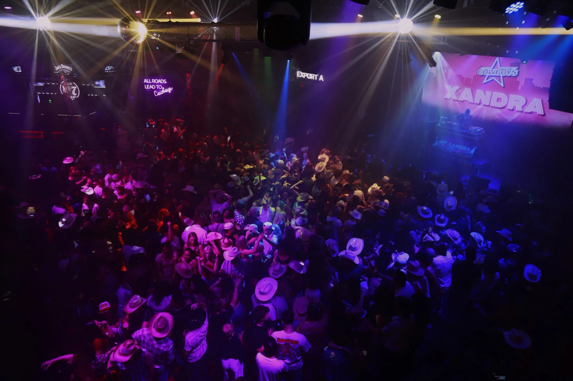 Crowded nightclub dance floor with cowboy hats under colourful stage lights and DJ booth.