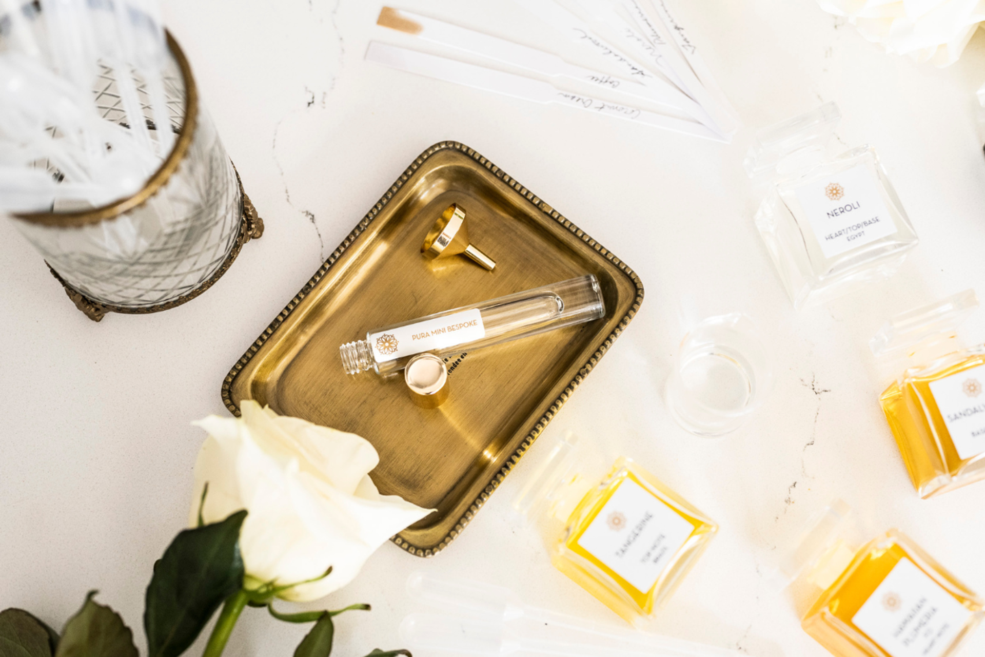 Perfume vial on a brass tray surrounded by scent bottles, strips, and a white rose.