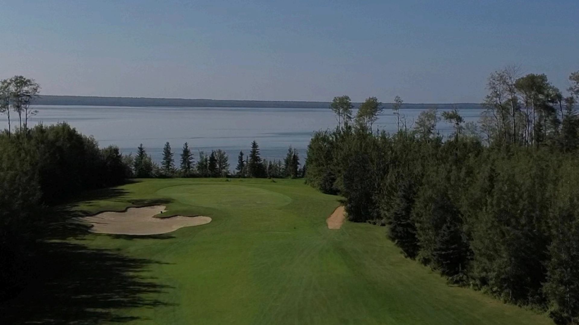 Golf course fairway and sand bunkers bordered by trees beside a large lake.