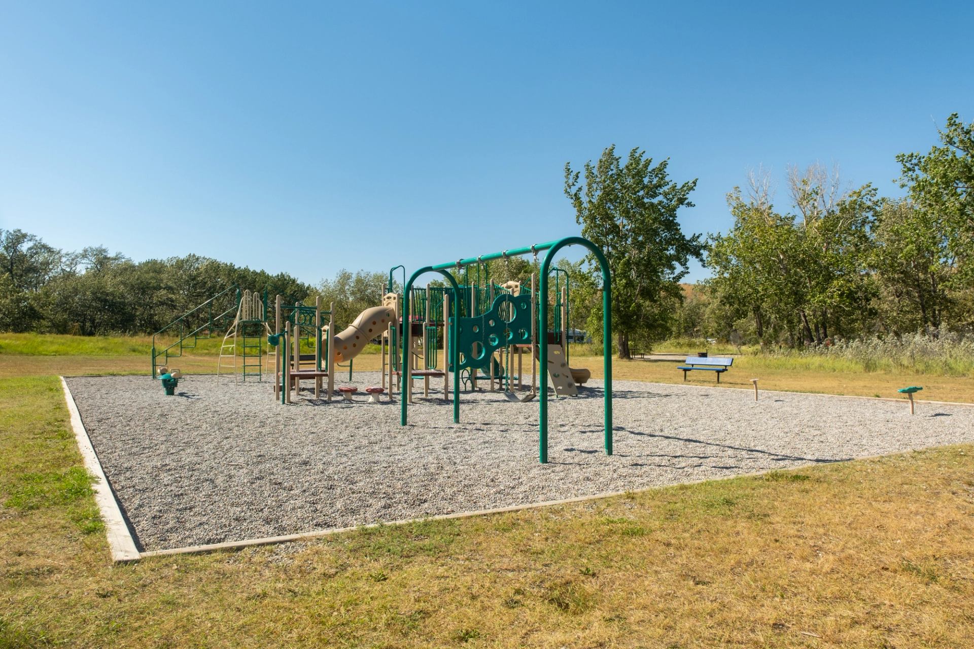 An outdoor playground with slides, swings, and climbing equipment on wood chips, surrounded by grass and trees under a clear blue sky.