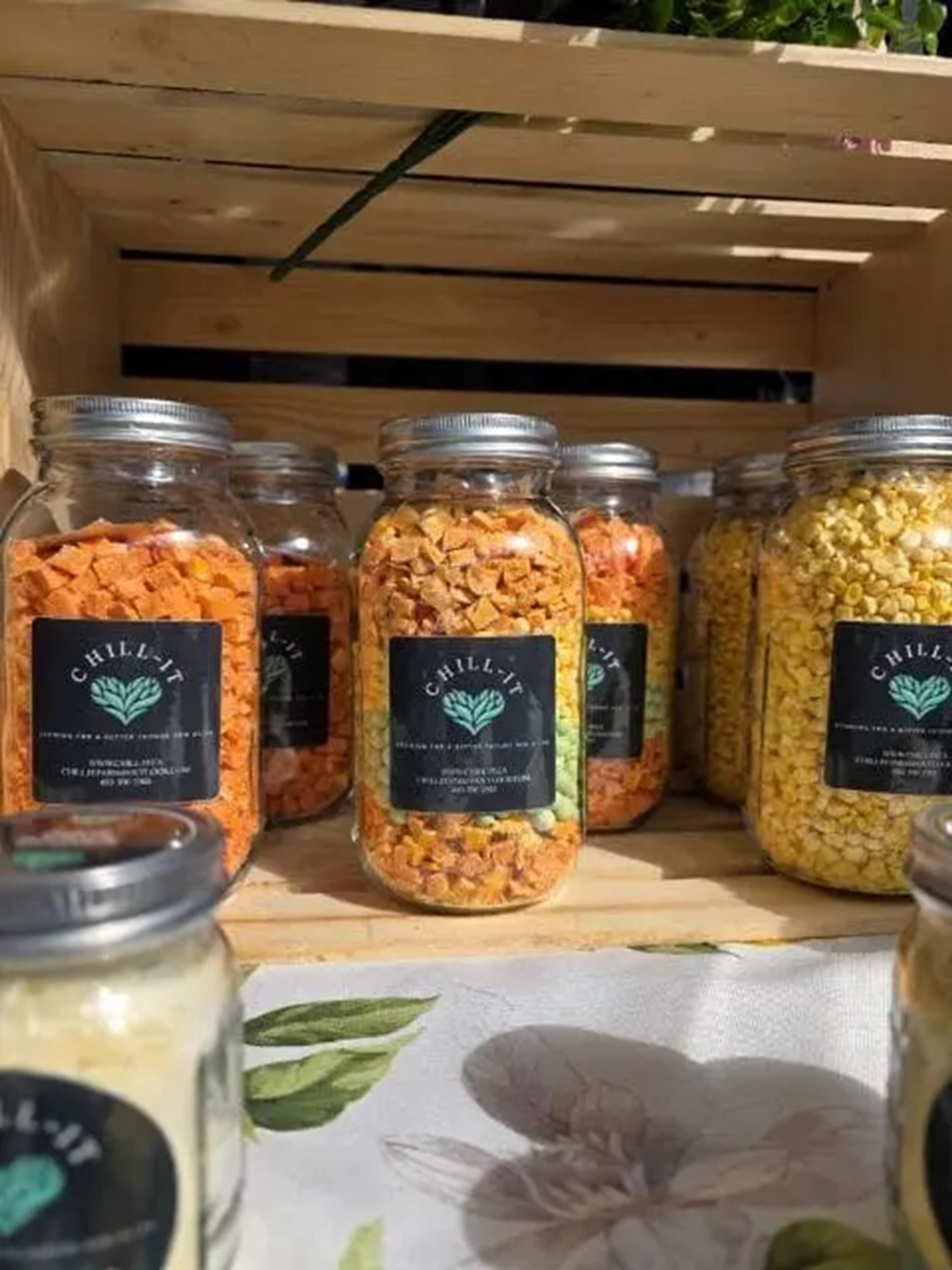 Glass jars of colourful dried vegetables with Chill-It labels displayed in a wooden crate.