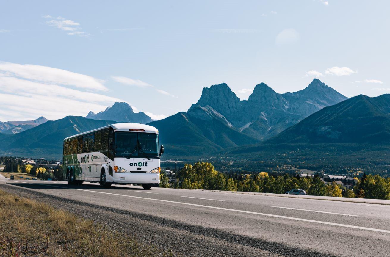 White coach bus on highway with mountain backdrop and clear blue sky.