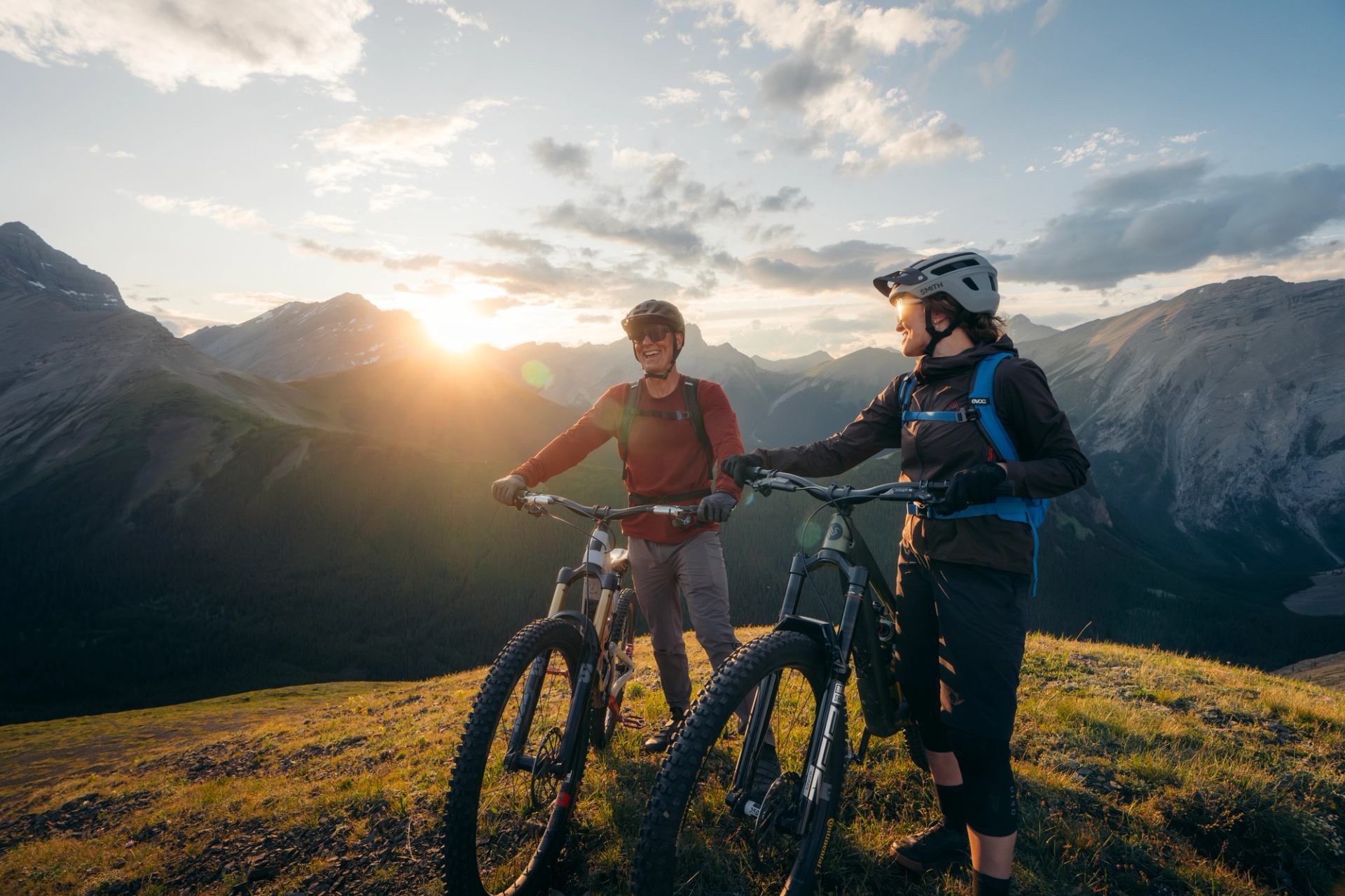 Two mountain bikers paused on a ridge at sunset with sweeping mountain views and dramatic skies.