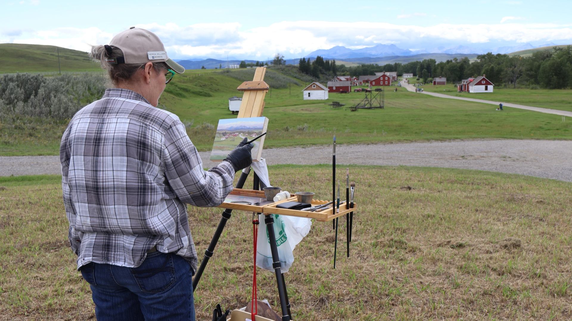 An artist paints a ranch landscape on an easel with historic buildings in the distance.