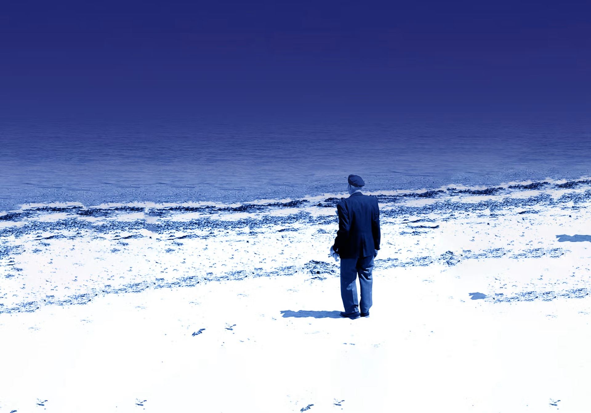 Person in dark clothing stands on bright beach facing calm blue ocean horizon.