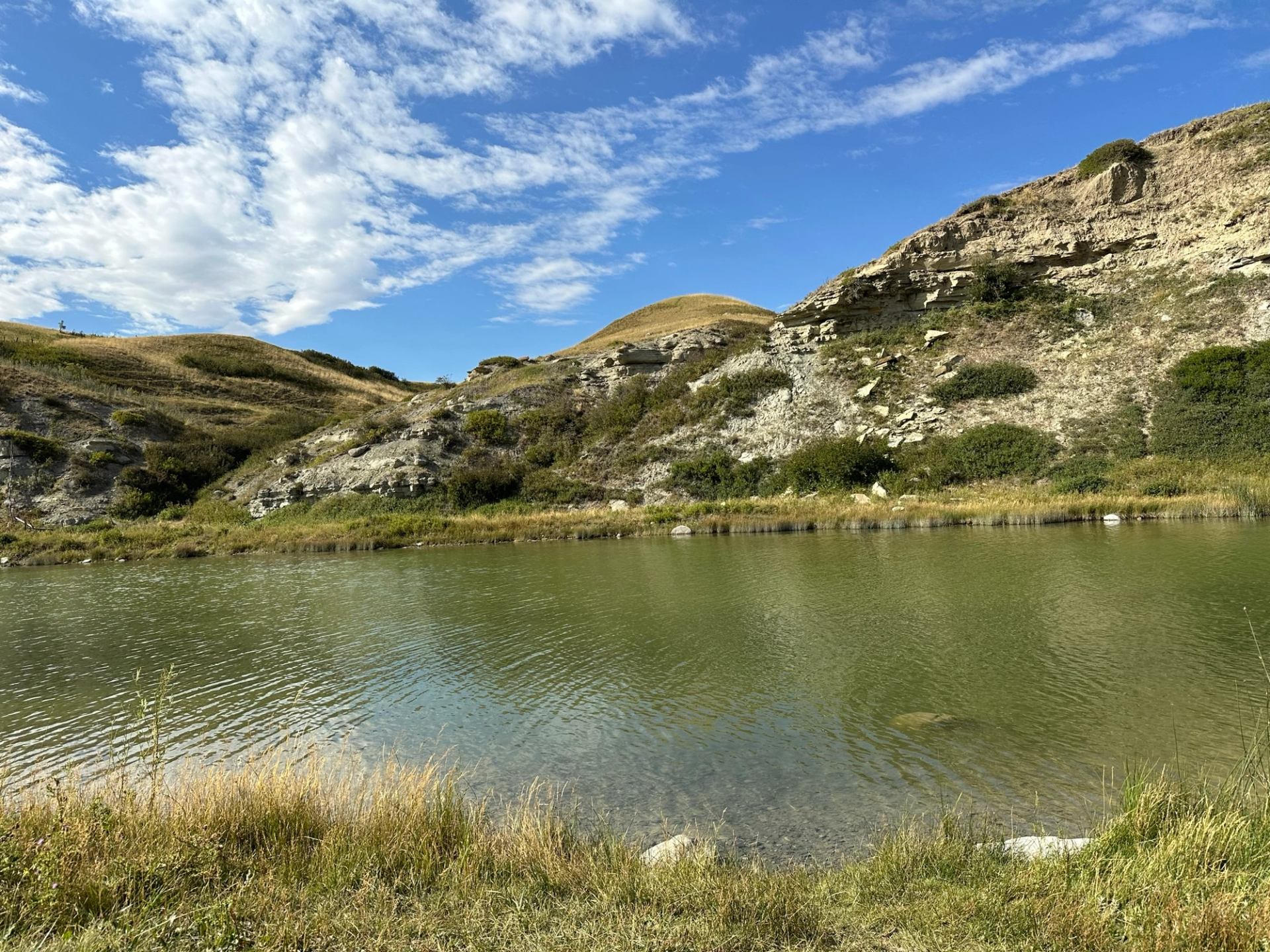 A calm river beside rocky coulee cliffs under a blue sky with scattered clouds.