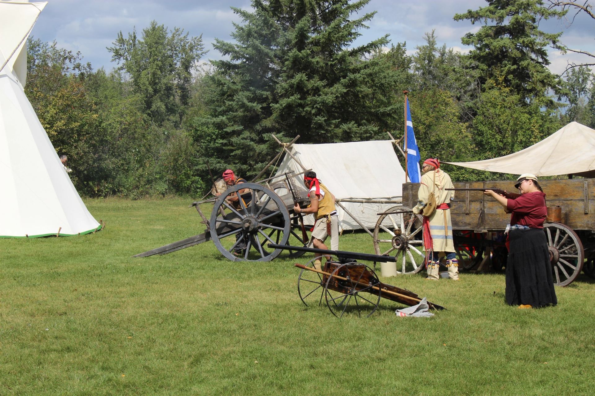 Historical re-enactors operate cannons and a rifle beside canvas tents and wooden wagons.