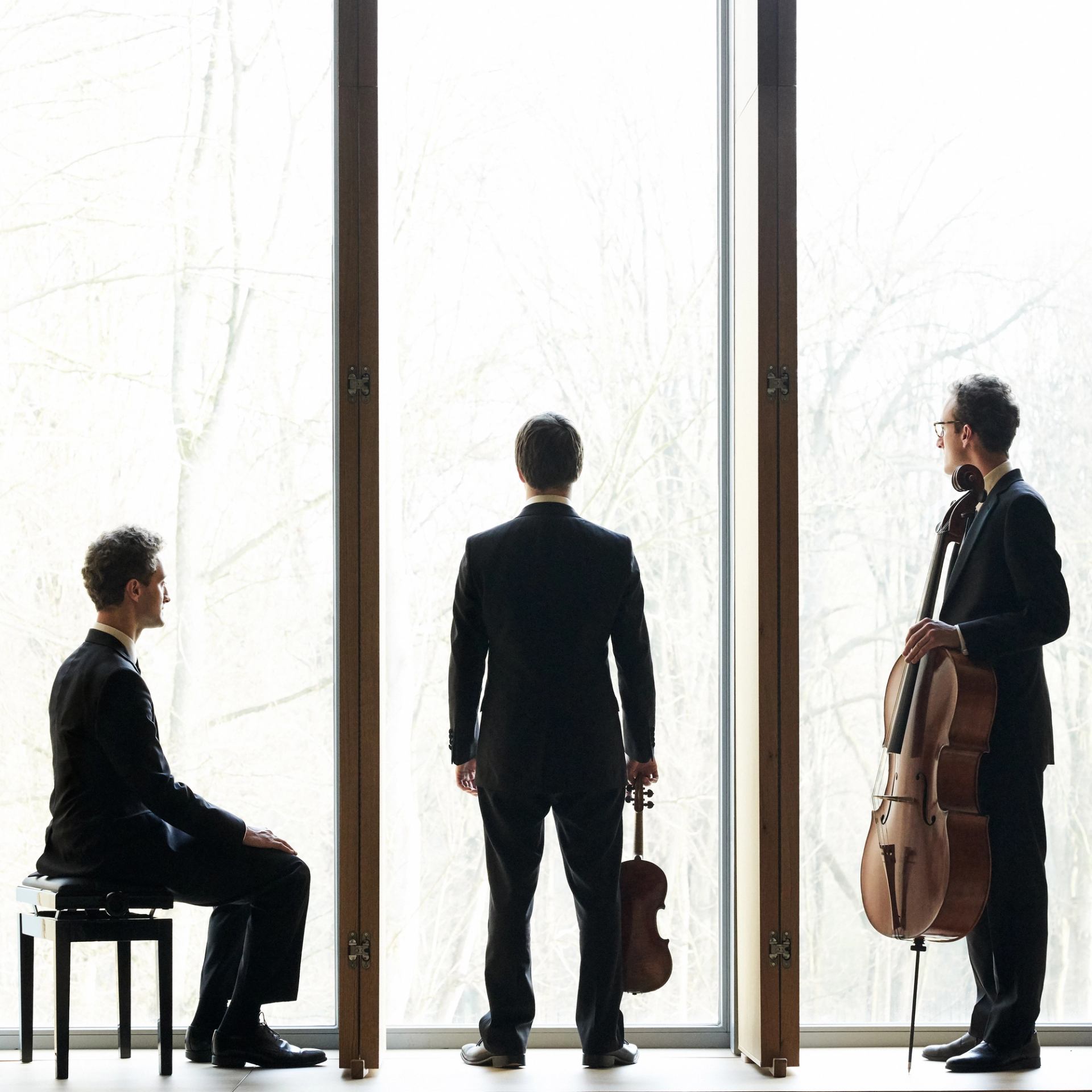 Three musicians in suits by tall window, holding violin and cello, facing outside.