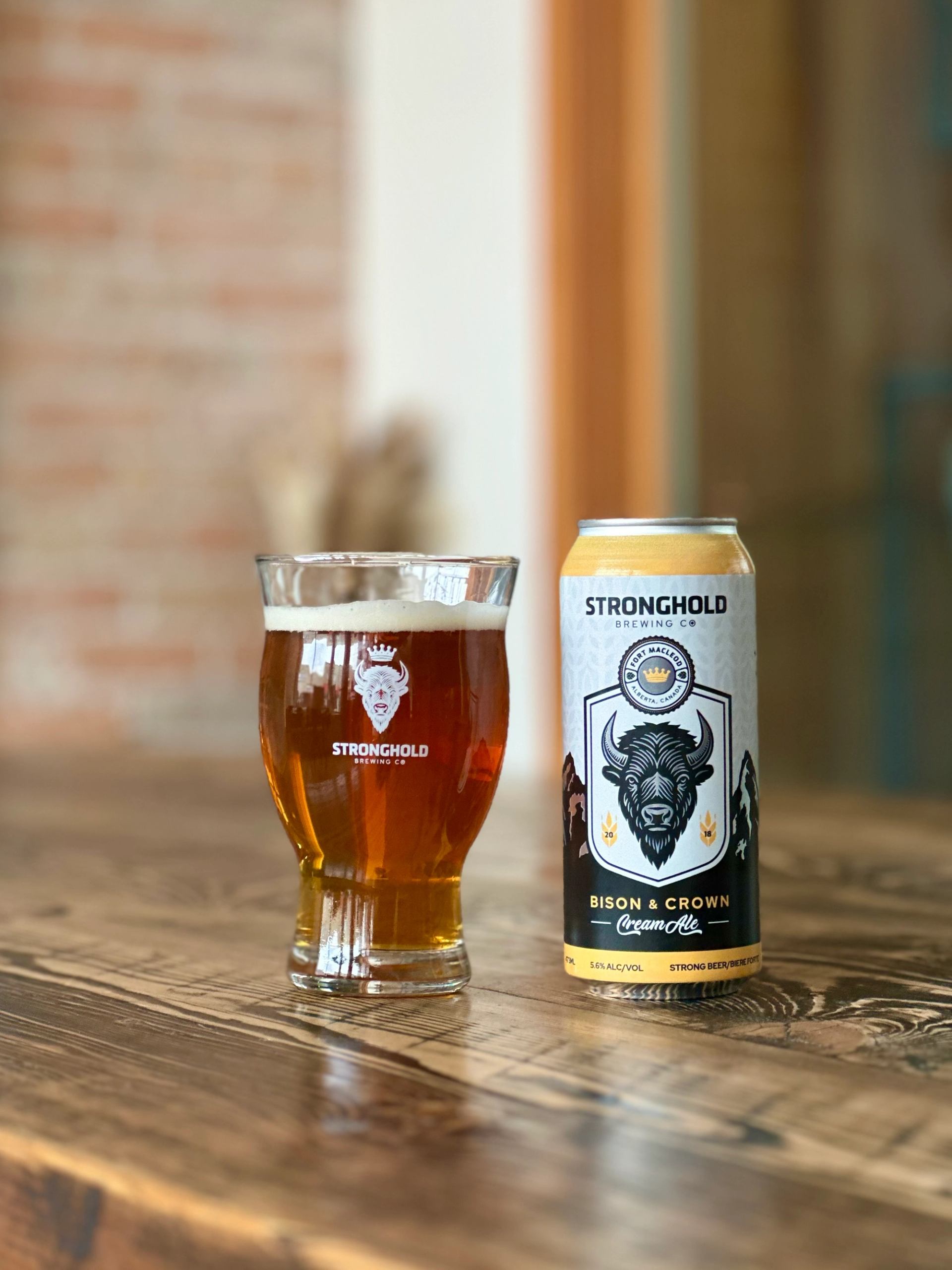 Glass of amber beer beside a can of Stronghold Bison & Crown Cream Ale on a wooden table.