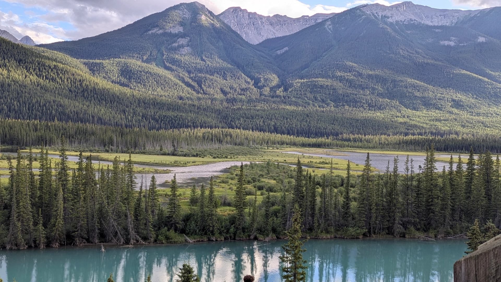 Turquoise river with forest, hills, and rocky mountains under cloudy sky.
