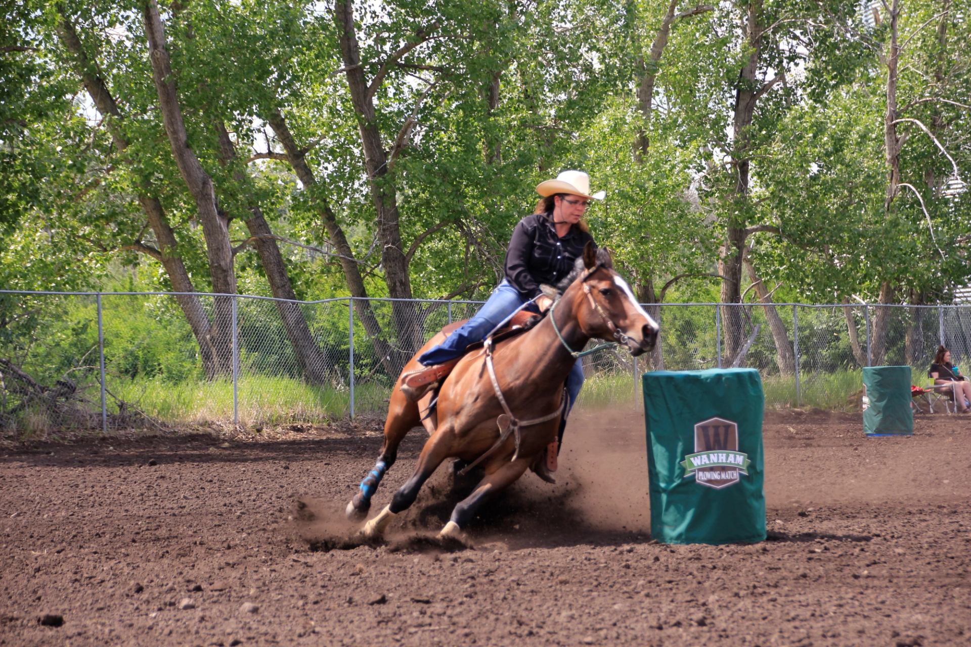 Barrel racer guiding a horse through a fast turn around a green barrel on a dirt arena.