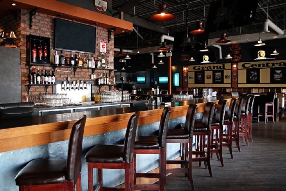 Rustic bar with brick walls, high chairs, shelves of bottles, and a large TV screen.
