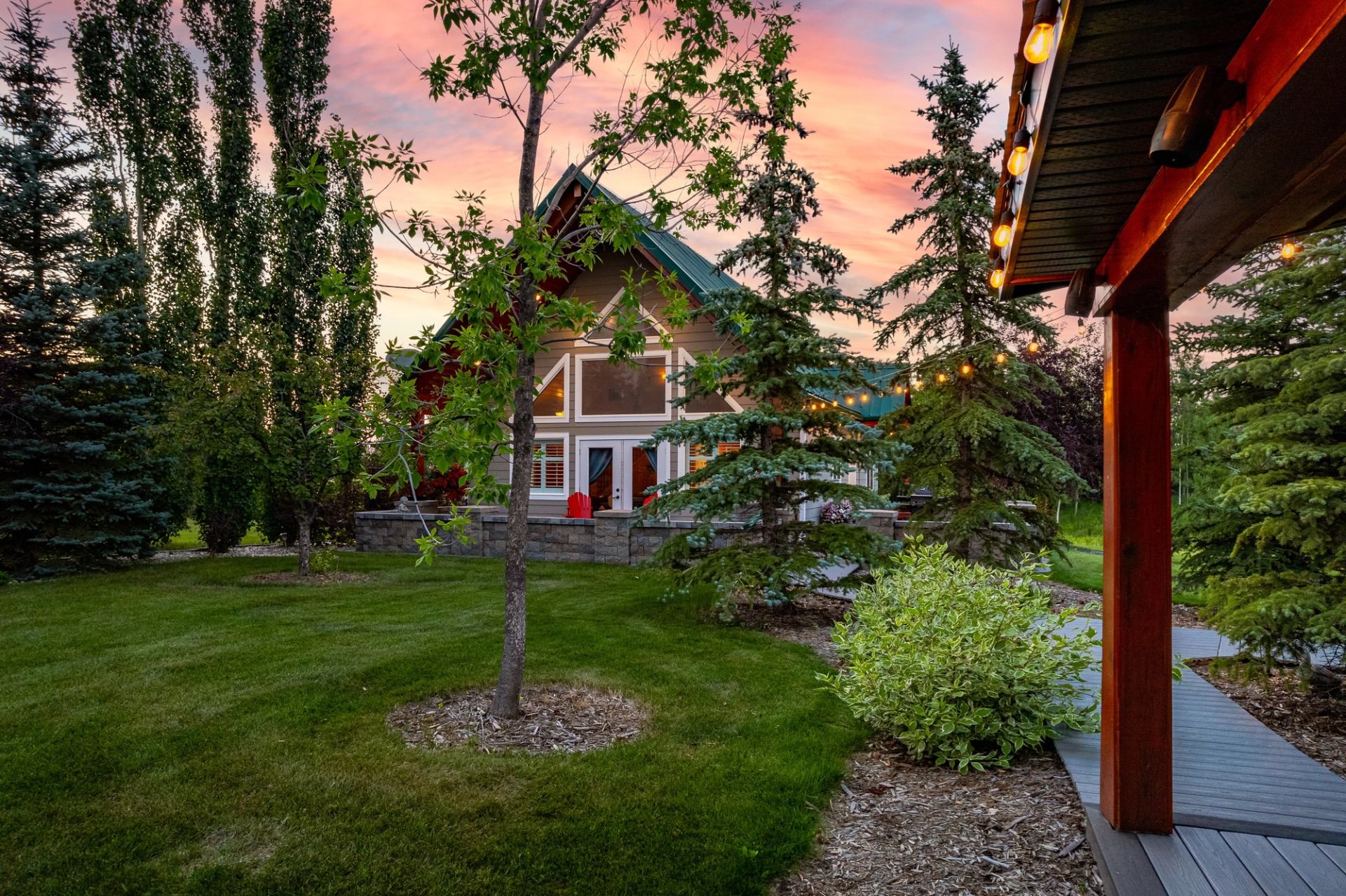 Cabin with green roof framed by trees and string lights at sunset in a landscaped yard.
