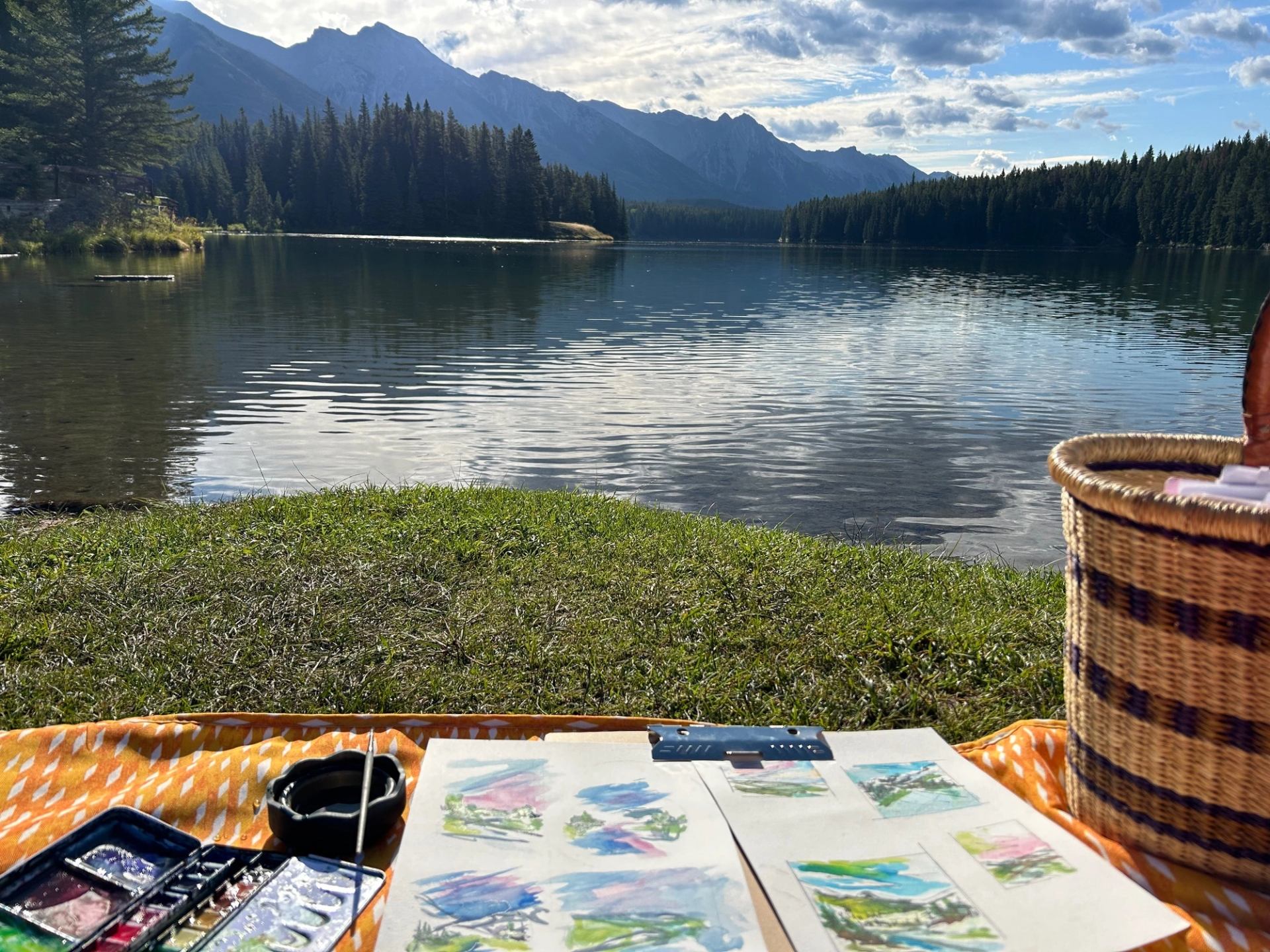 Mountain lake with fir trees, cloudy sky, and painting supplies on a blanket by shore.