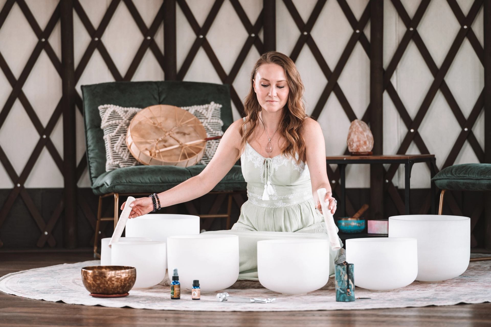 Person plays white singing bowls with mallets while seated on a patterned rug.
