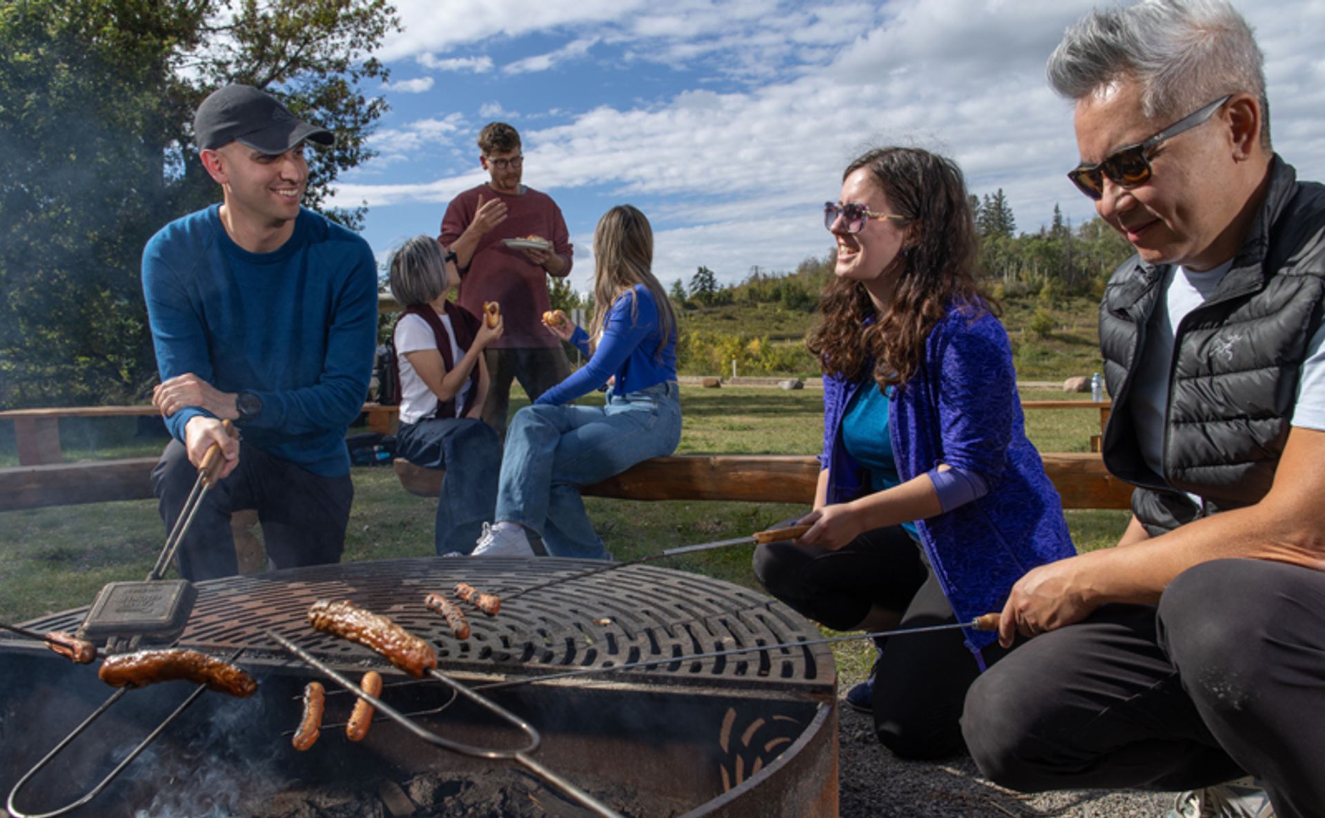 People roast sausages over a circular outdoor grill in a grassy park setting.