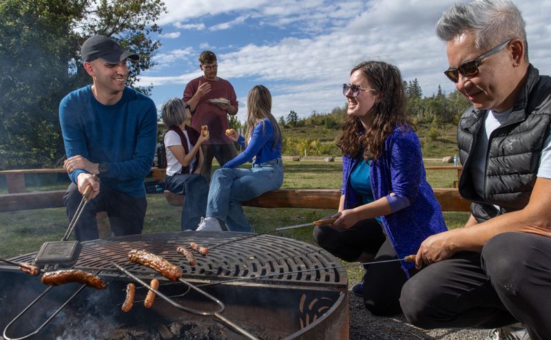 People roast sausages over a circular outdoor grill in a grassy park setting.