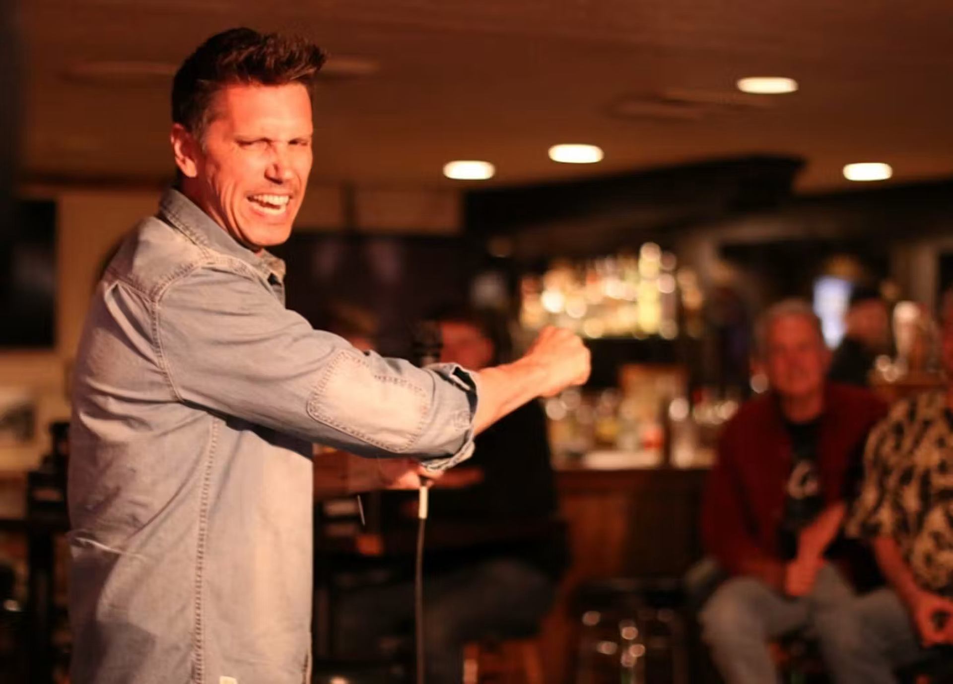 Comedian in denim shirt gestures on stage with microphone before seated audience.
