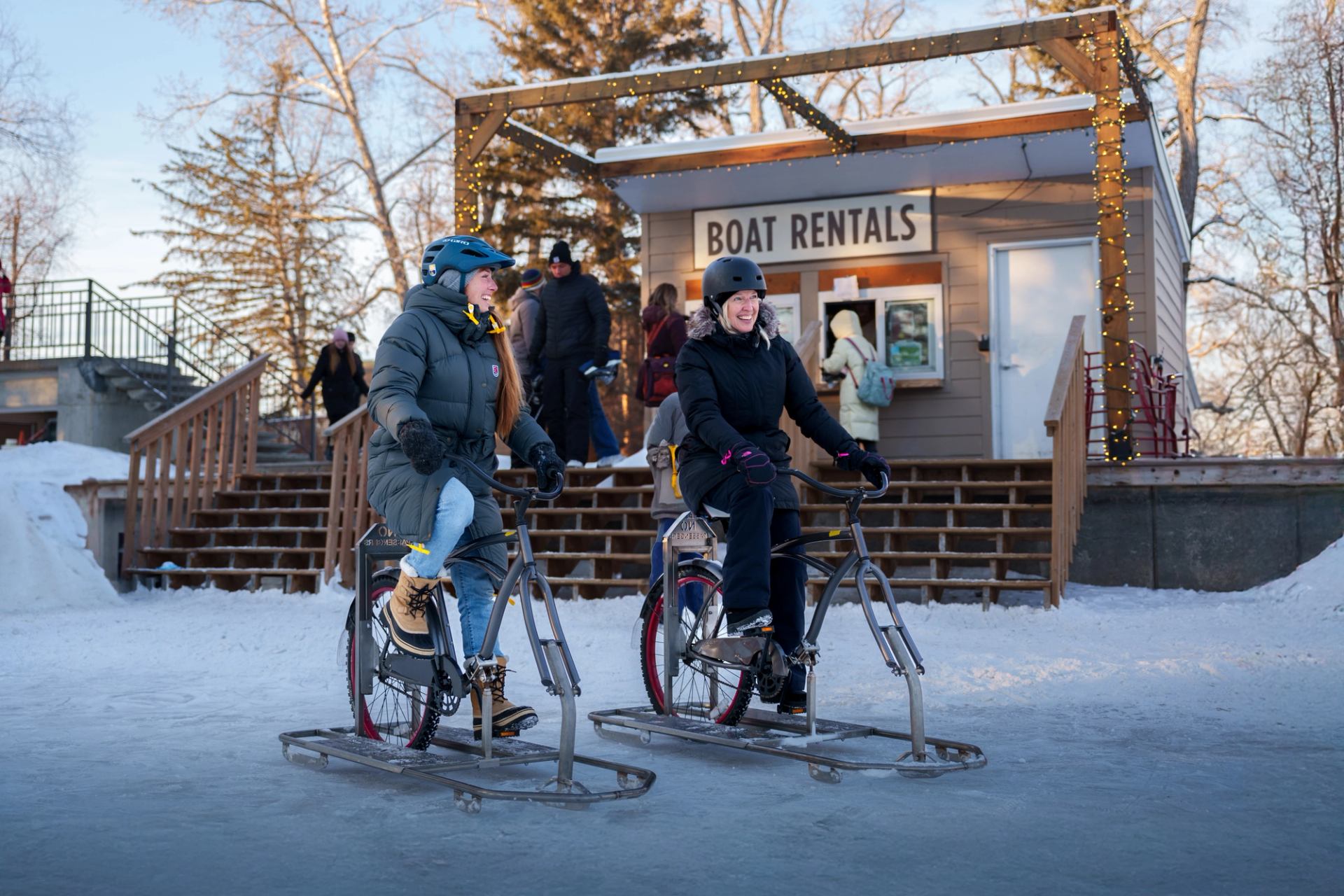 Two people riding ice bikes near a boat rental hut at Bowness Park in winter.