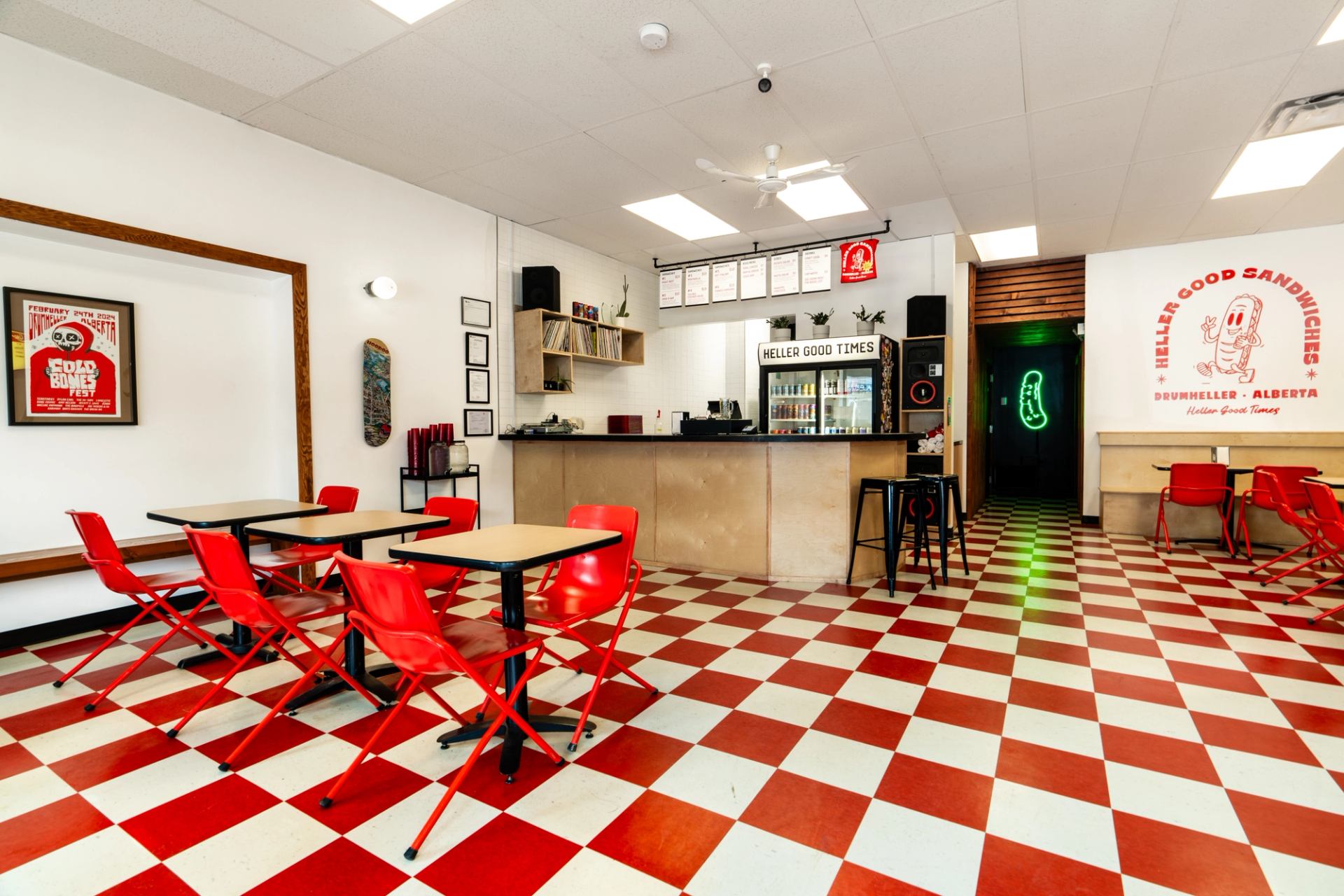 A casual sandwich shop interior with a red and white checkered floor, red chairs, a wooden counter, and a neon hot dog sign.