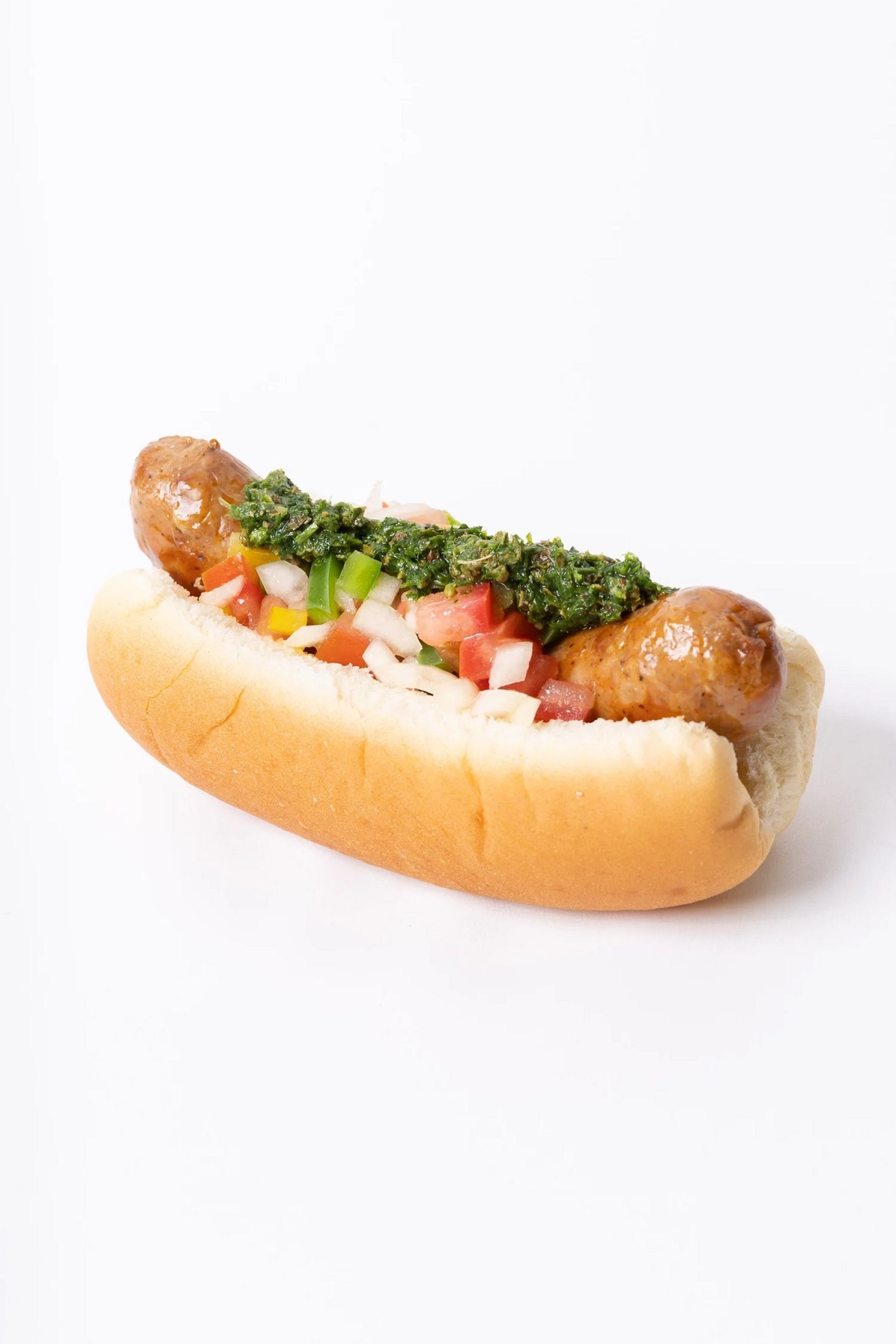 Sausage in a bun topped with diced vegetables and green herb sauce on white background.