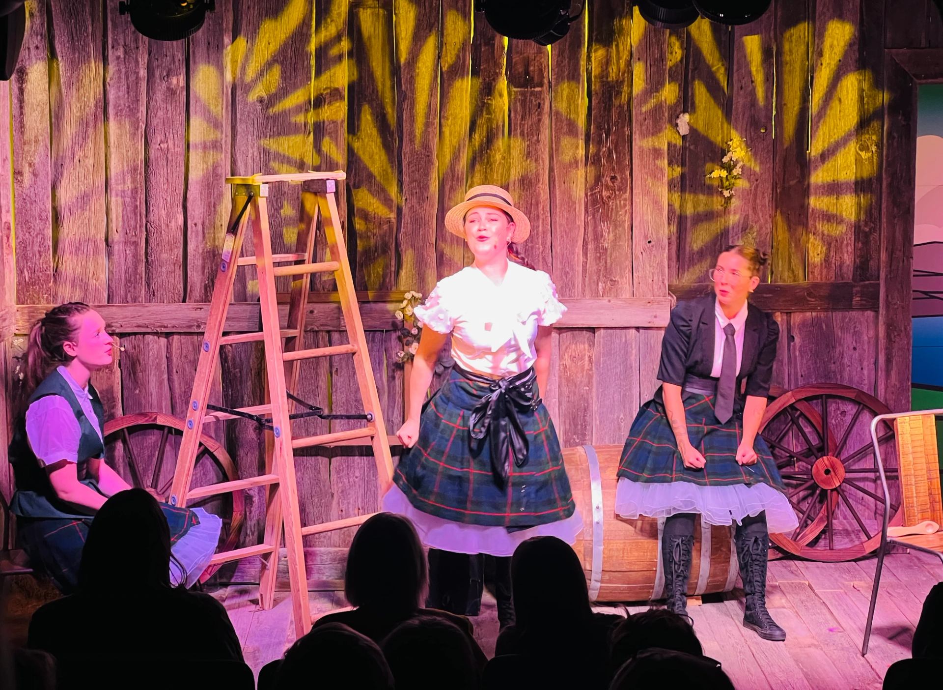 Three performers in kilts act on a wooden stage with barrels, wagon wheels, and theatrical lighting.