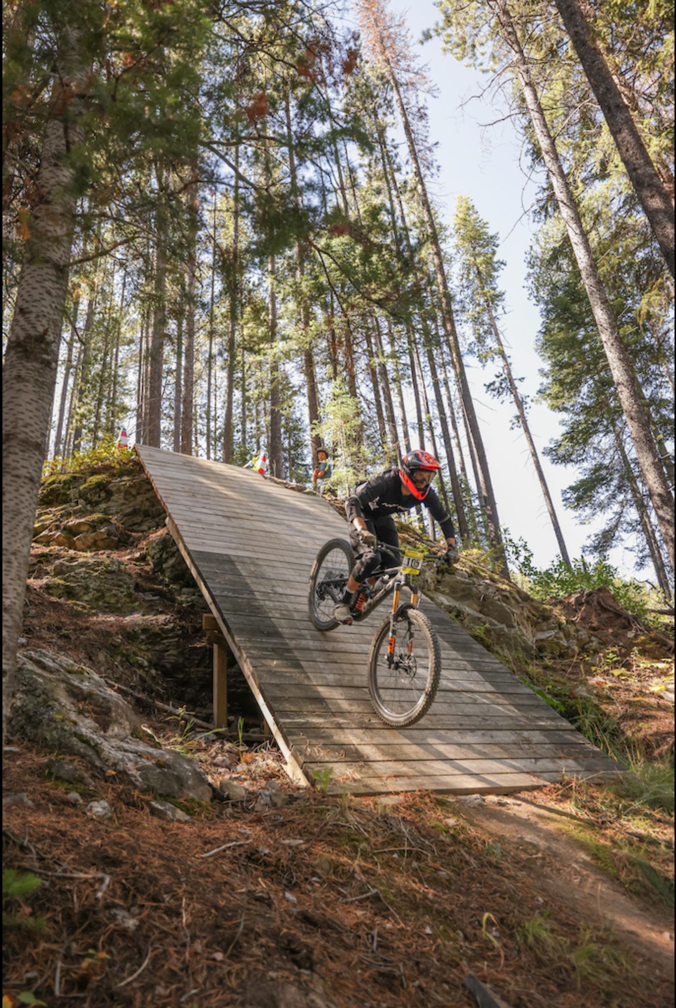 Mountain biker rides a wooden ramp on a trail through a forest.