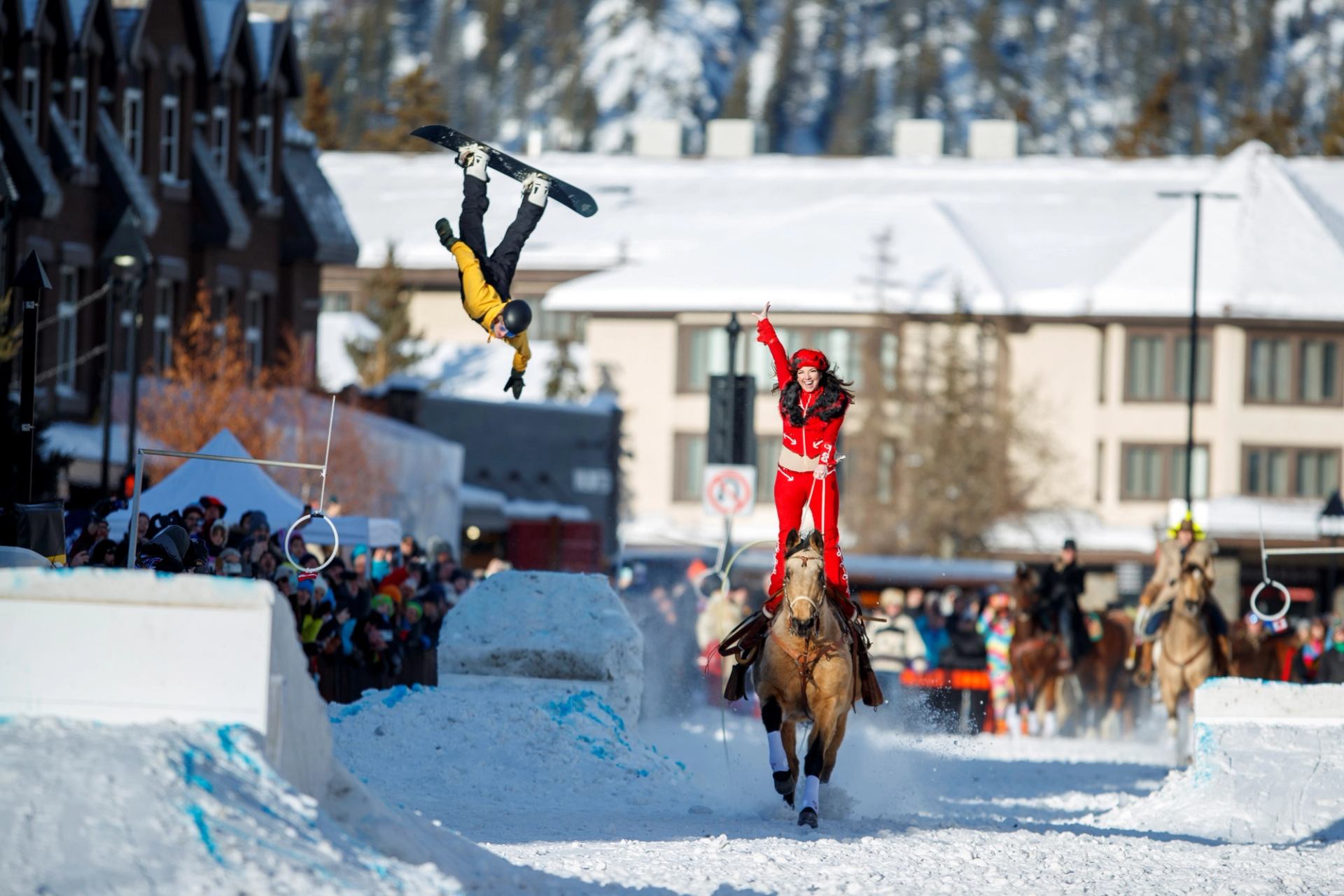 Winter sports scene with horse, rider, and flying snowboarder.